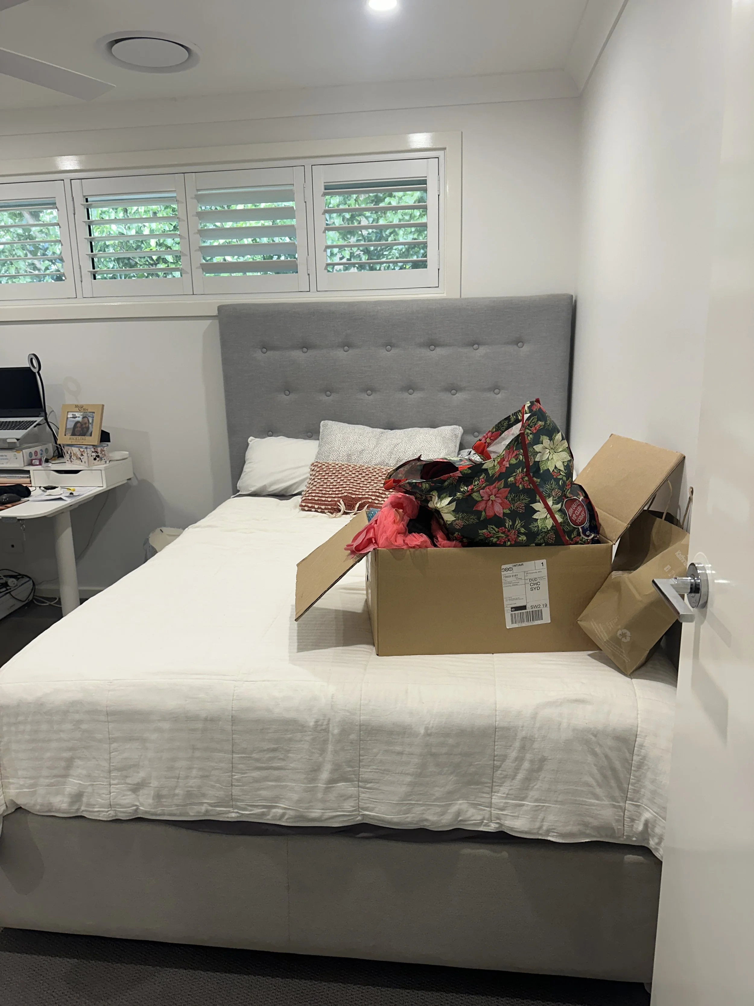 An unmade bed with a gray headboard and a white bedspread. There is a cardboard box with red and black items and a floral tote bag on the bed. To the left, a small desk holds a laptop, photo frame, and various items. Behind, there are three horizonta