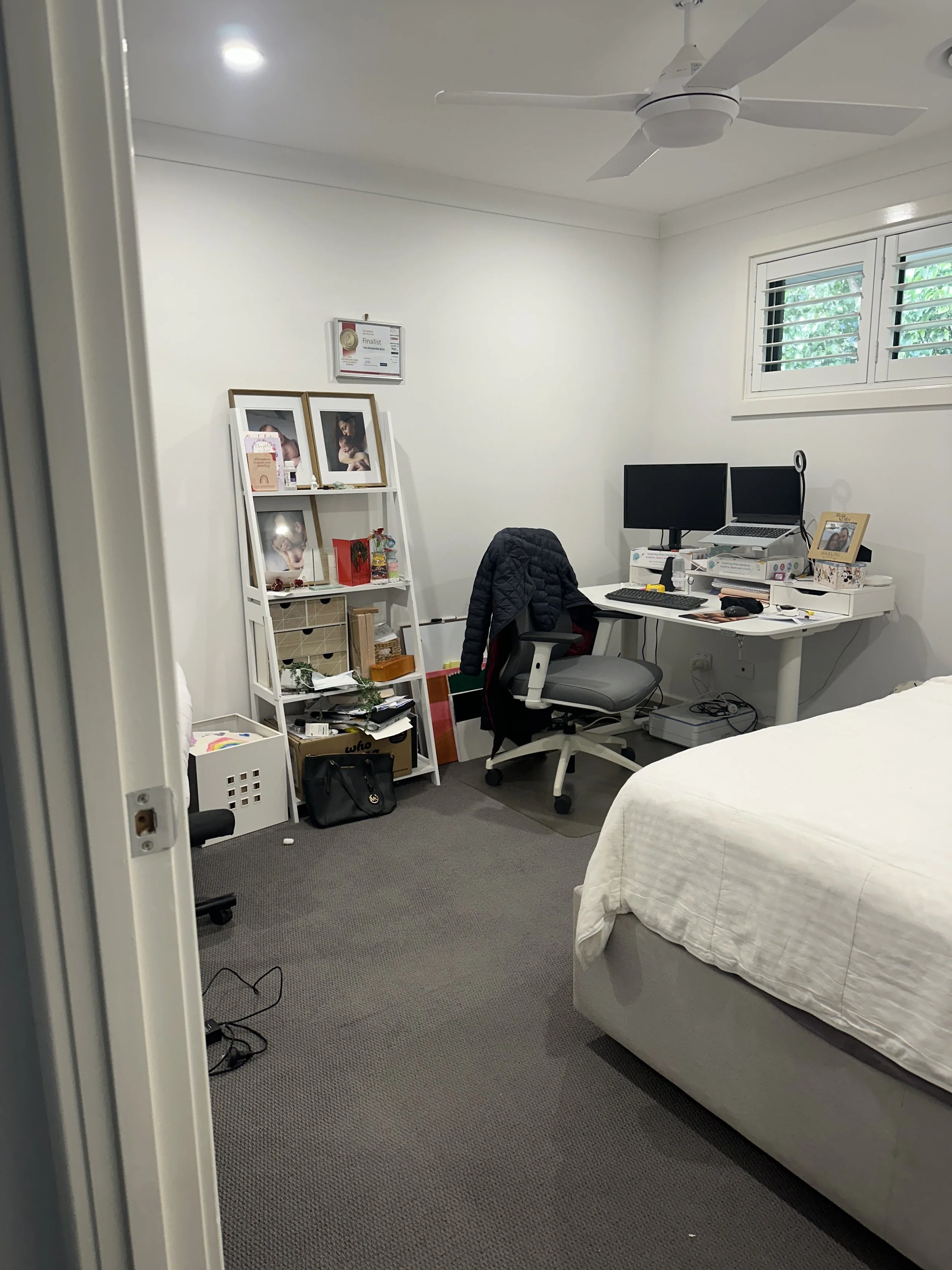 A bedroom with a white bed, a work desk with two monitors, a laptop, and various office supplies. A white bookshelf with framed photos and decorations is in the corner. A ceiling fan is above, and a window with louvered blinds is on the wall.