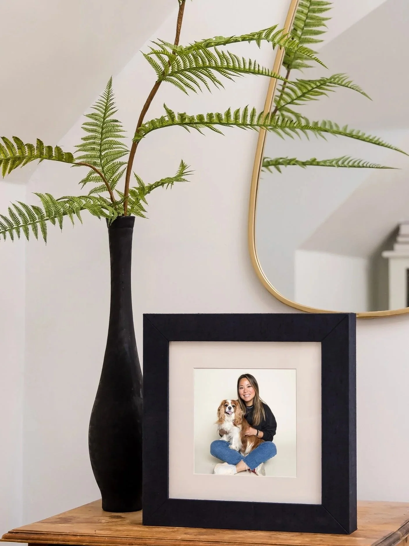 A framed photograph of a woman with long hair smiling and holding a small brown and white dog, placed on a wooden surface with a large black vase holding green fern leaves and a gold-framed mirror in the background.