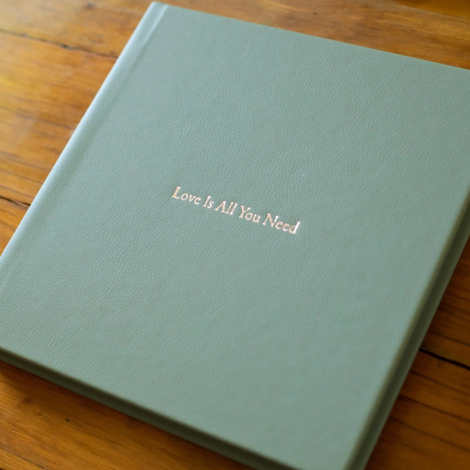 A hardcover book with a textured, soft green cover, resting on a wooden surface. The title, partially visible, reads "Love Is All You Need" in small, gold lettering.