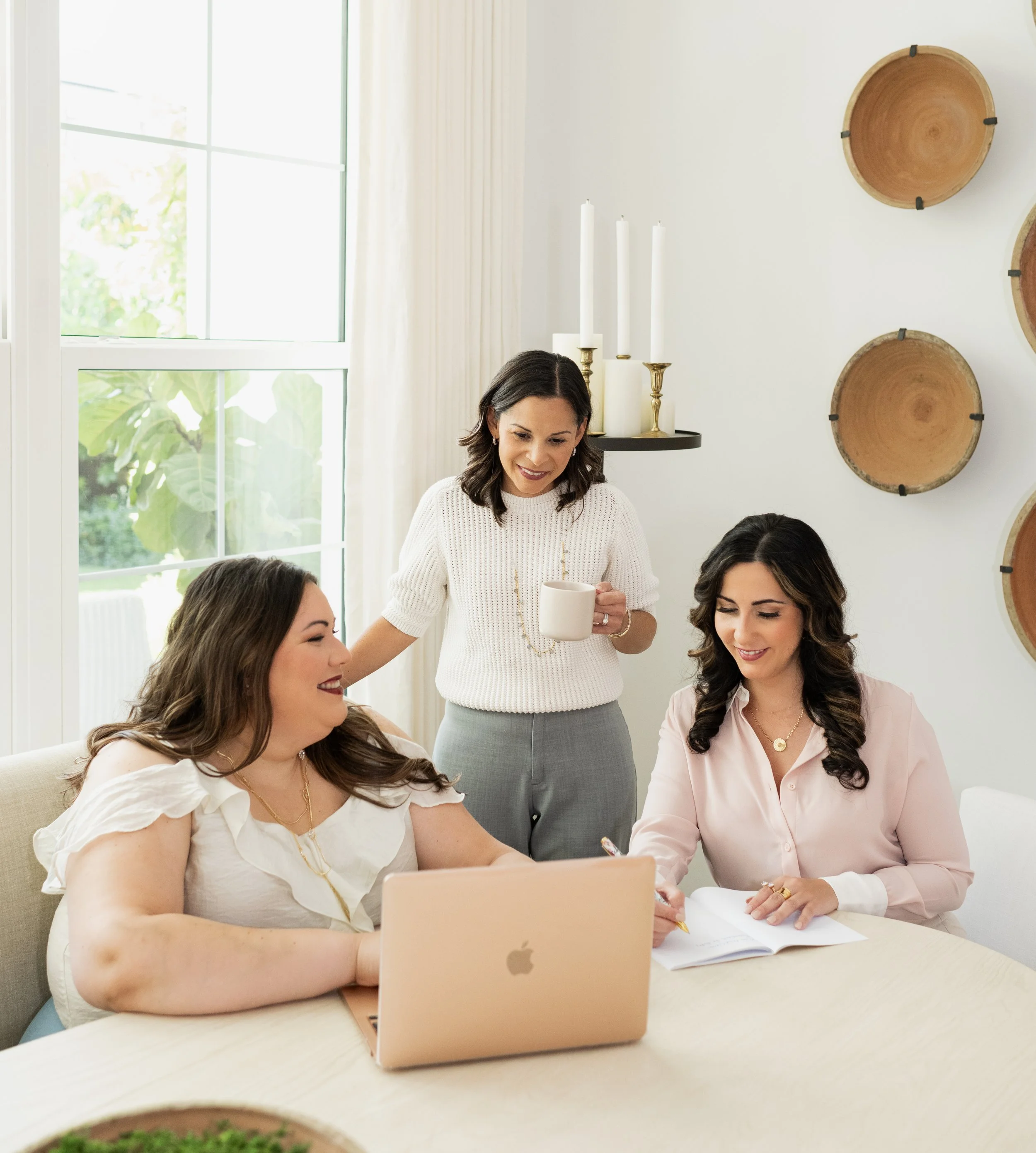 Three women working together at a table in a bright, airy room. One woman is sitting with a laptop, another is writing in a notebook, and the third is standing and holding a mug. The room has large windows and decorative plates on the wall.