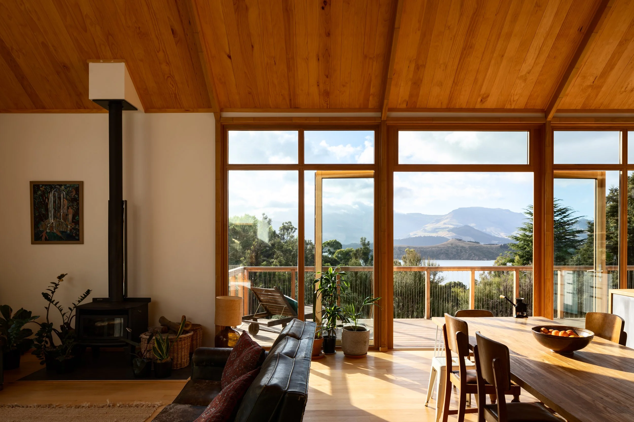 Open-plan living and dining area with timber-lined vaulted ceiling, wood burner and large windows framing the harbour views.