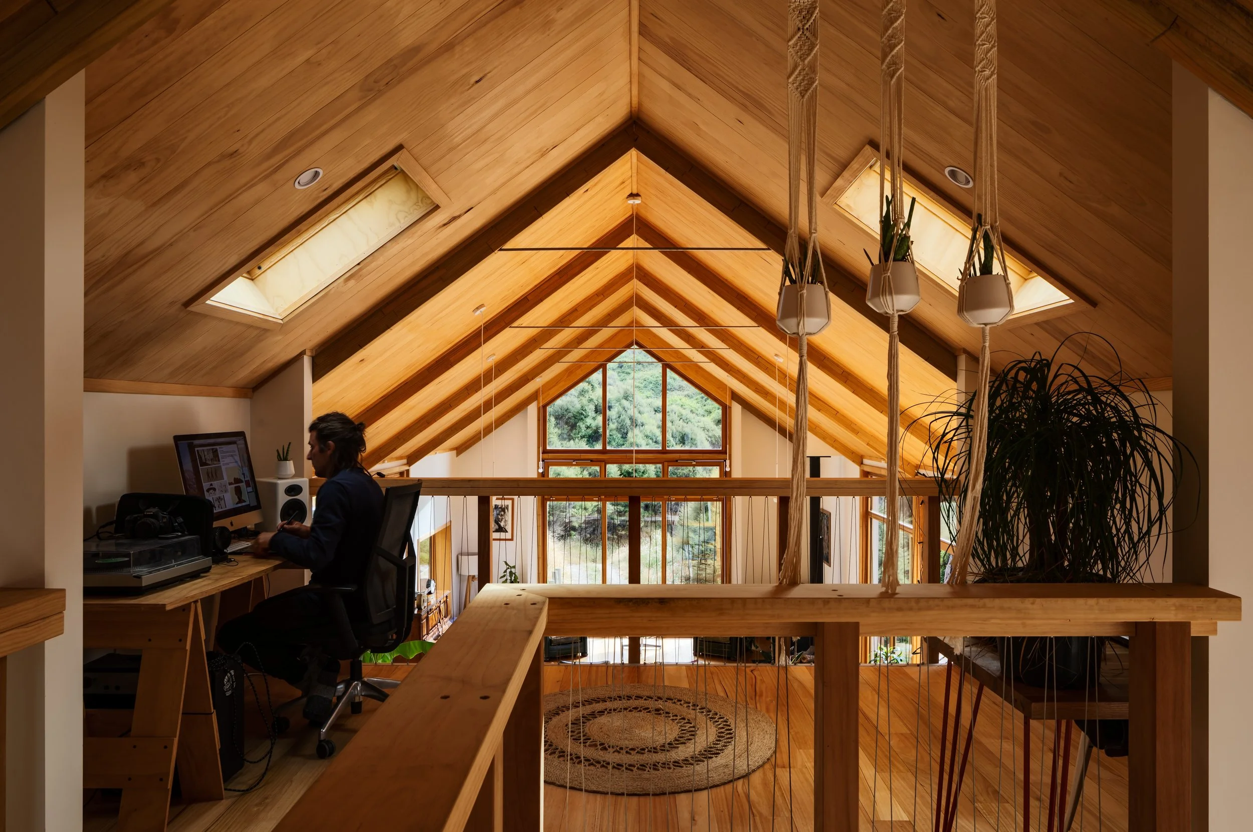 Mezzanine workspace beneath a timber gabled roof overlooking the double-height living area, with skylights and hanging plants.