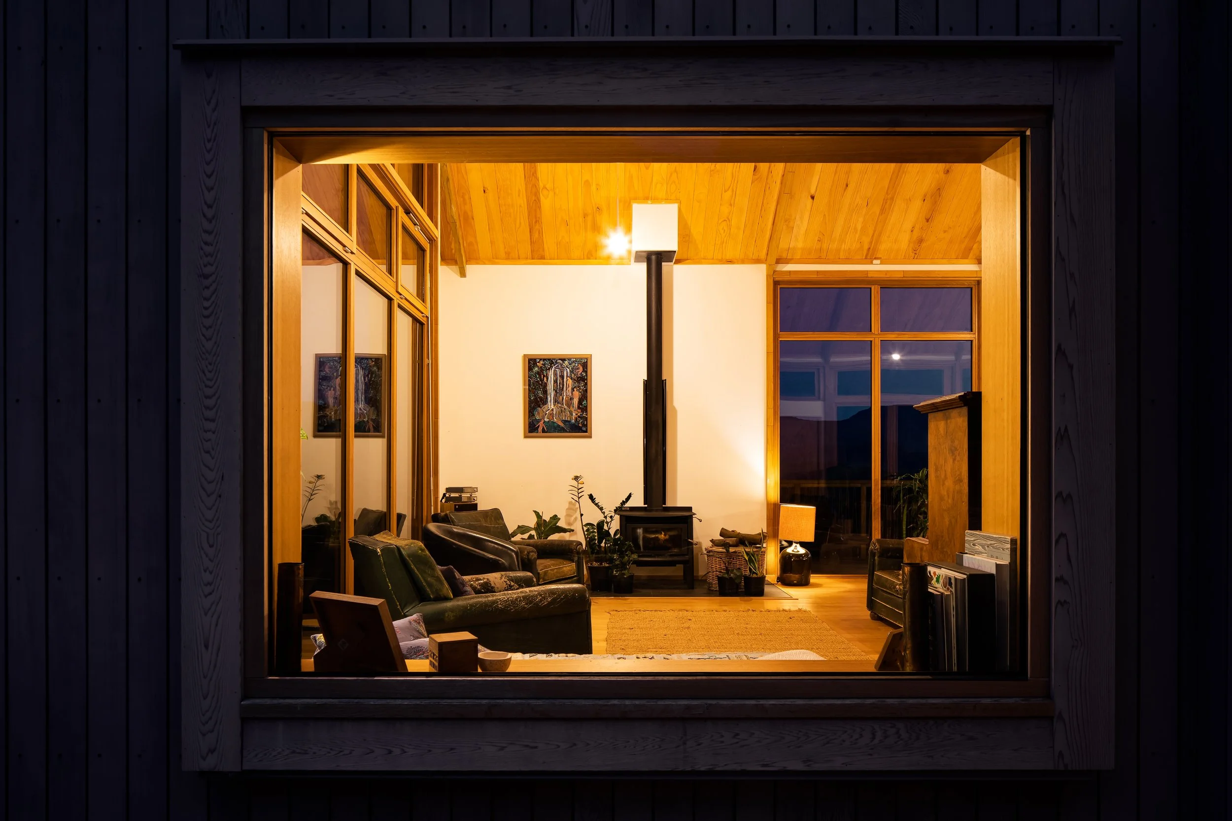 Warmly lit living room viewed through a large exterior window at dusk, showing the timber interior and wood burner.