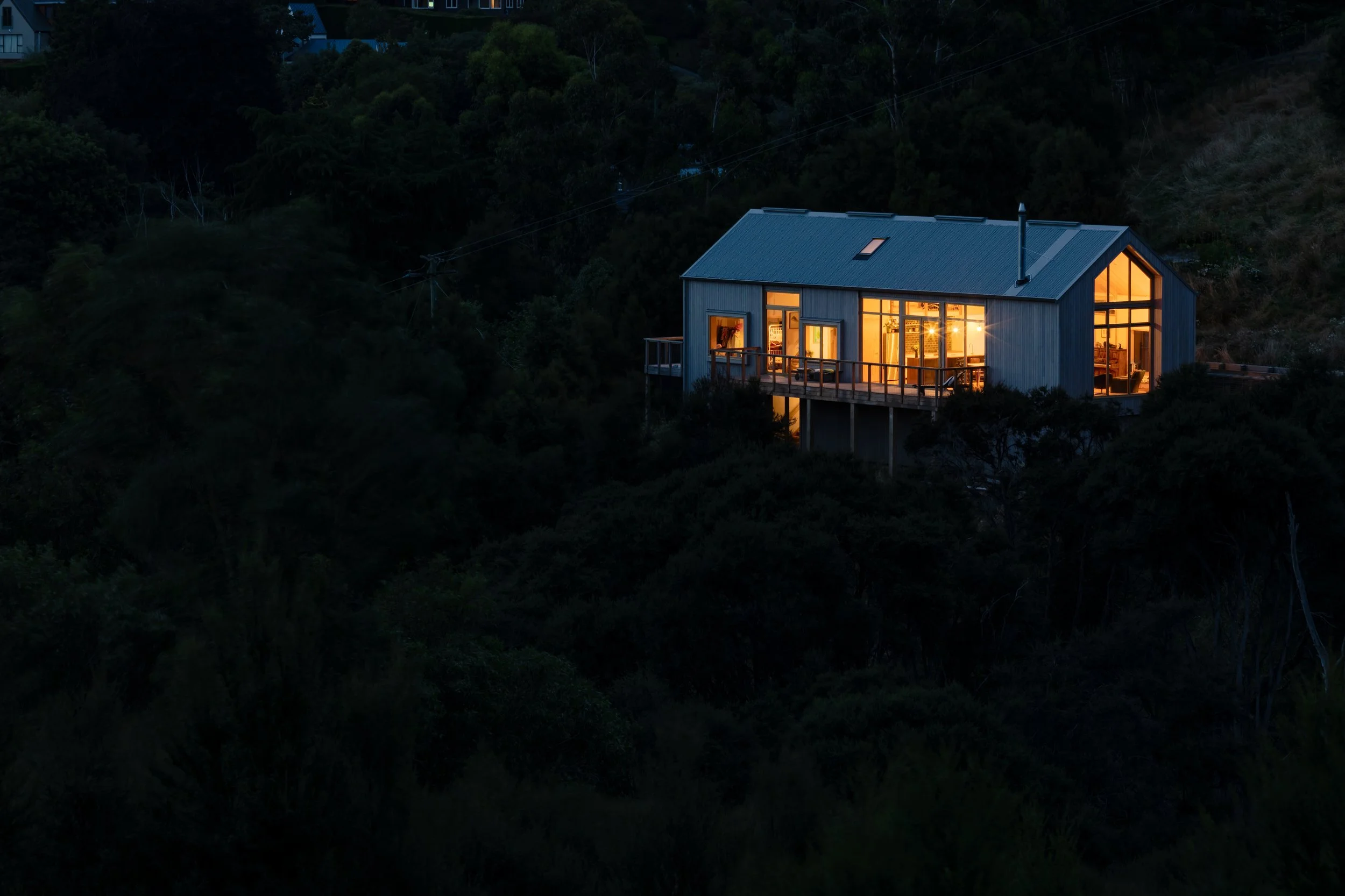 Timber-clad gabled house glowing from within at dusk, surrounded by vegetation on the Governors Bay hillside.
