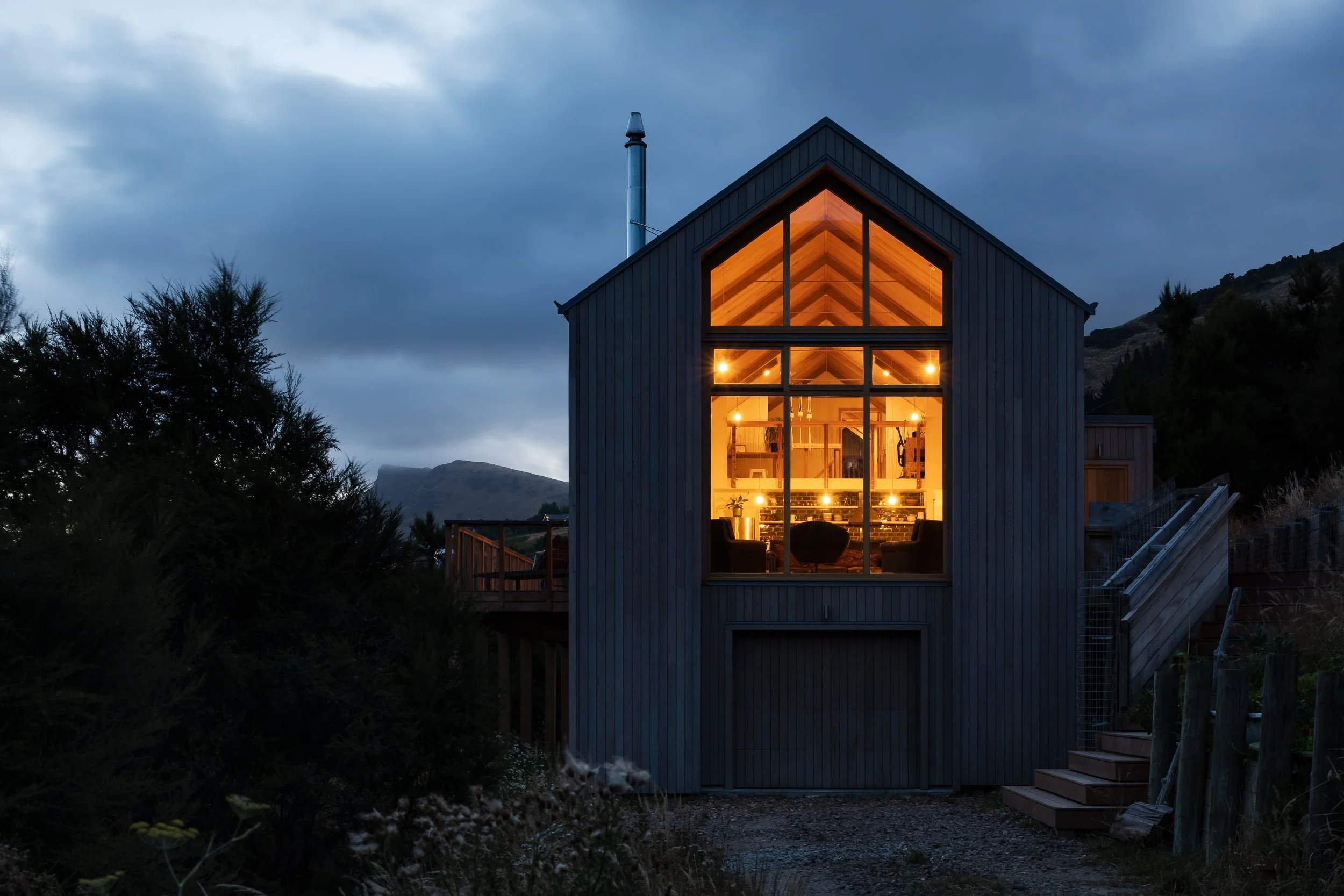 Timber-clad gabled house glowing from within at dusk, surrounded by vegetation on the Governors Bay hillside.