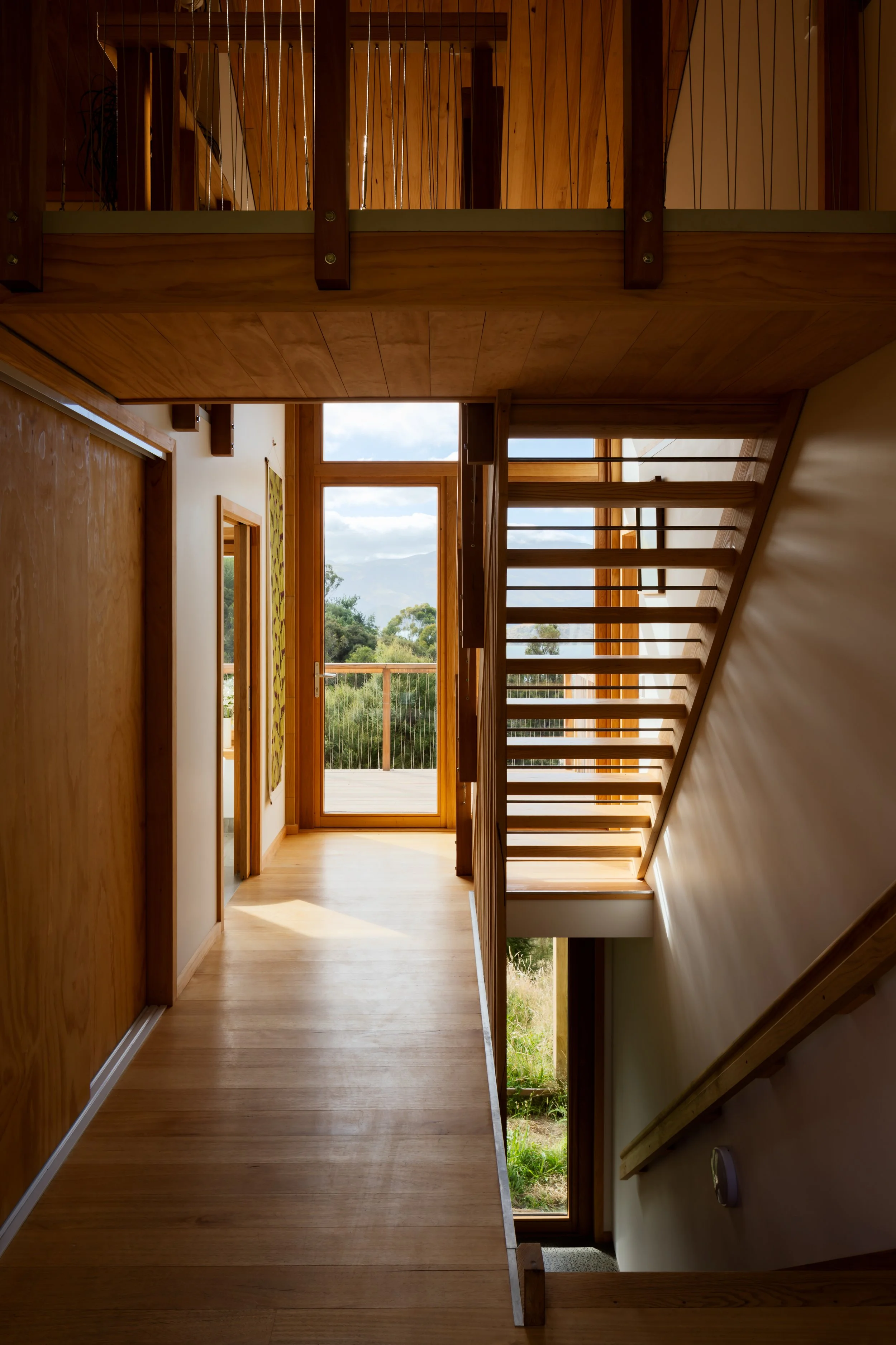 Timber-lined hallway with open-riser staircase, mezzanine and glazed door opening onto the deck.