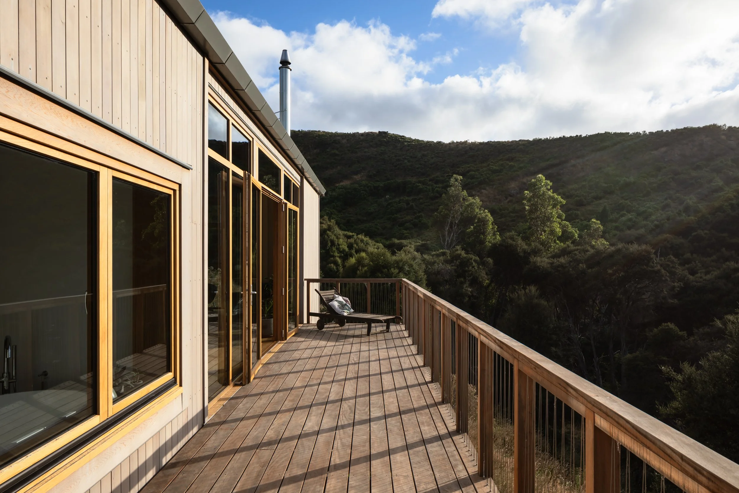 Timber-clad house with glazed doors opening onto a raised deck overlooking the surrounding native vegetation and hills.