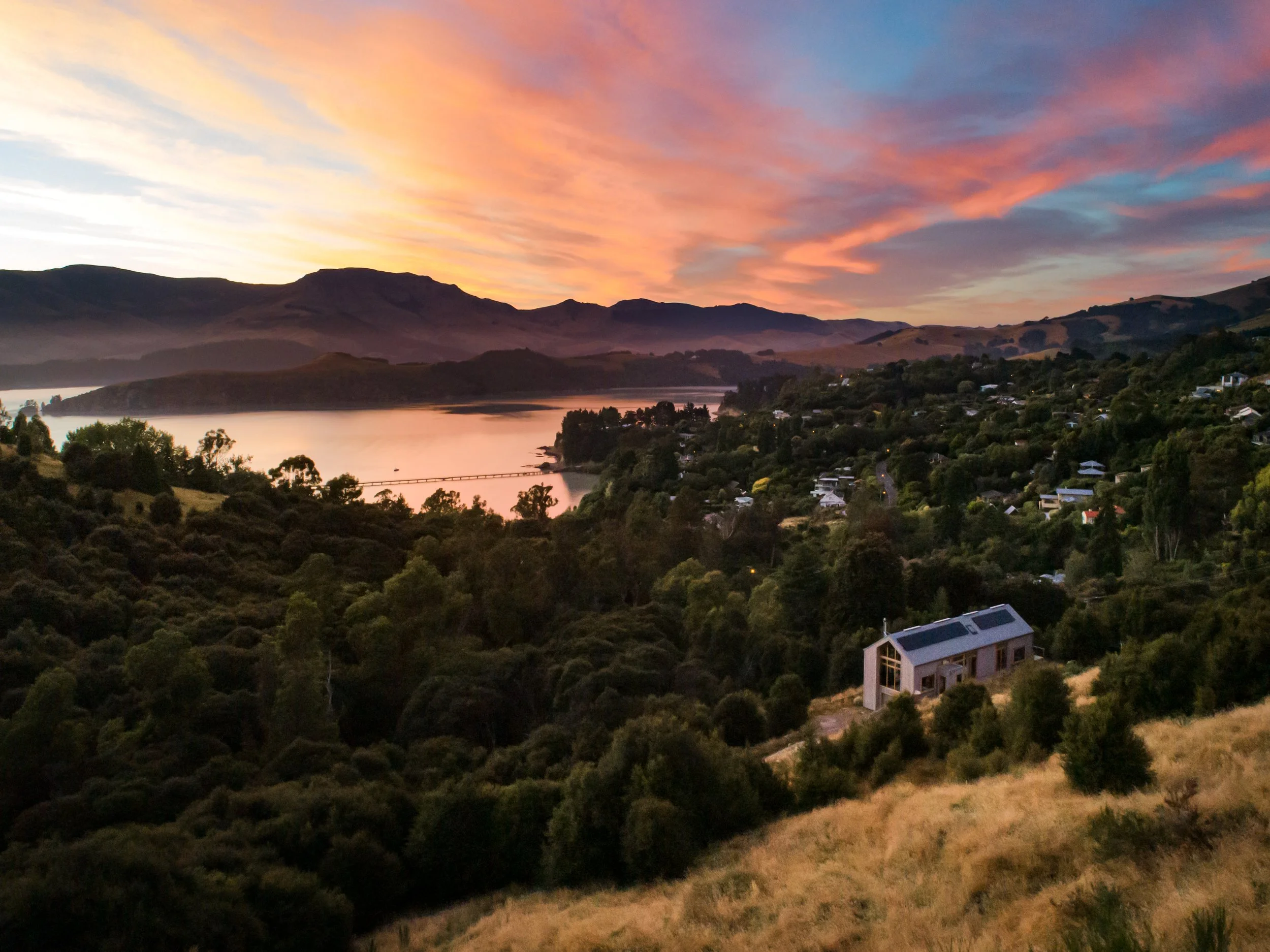 Aerial view of the timber Governors Bay house nestled in dense vegetation above Lyttelton Harbour at sunset.