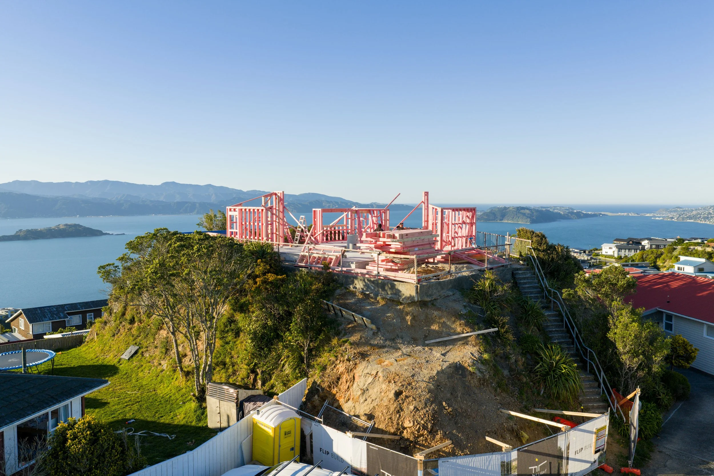 Construction site on a hillside overlooking a body of water with mountains in the background. The structure is in framing stage with pink lumber and has stairs, fencing, and construction equipment around.