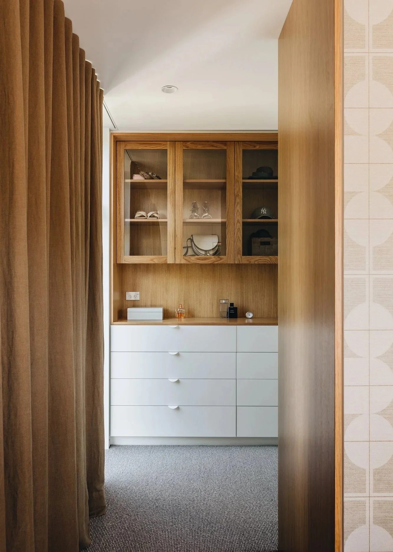 Bedroom closet with wooden shelves and white drawers, decorated with personal items and hats, seen through a doorway with brown curtains.