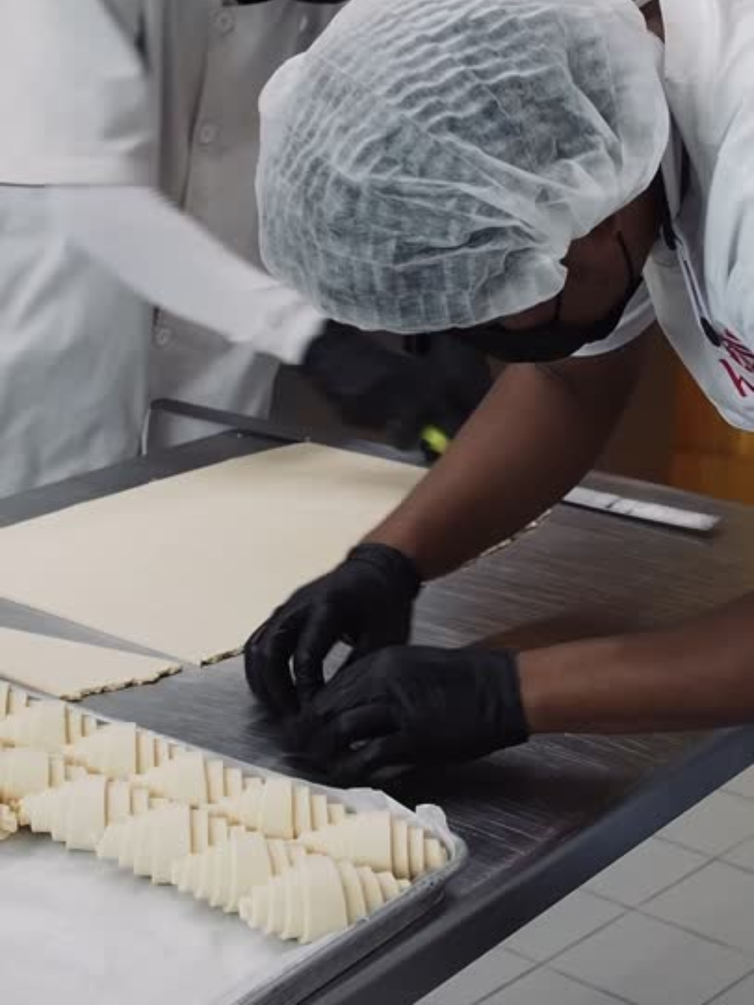 Person wearing black gloves and a hairnet is preparing a sheet of dough or cheese on a stainless steel table in a commercial kitchen or bakery.