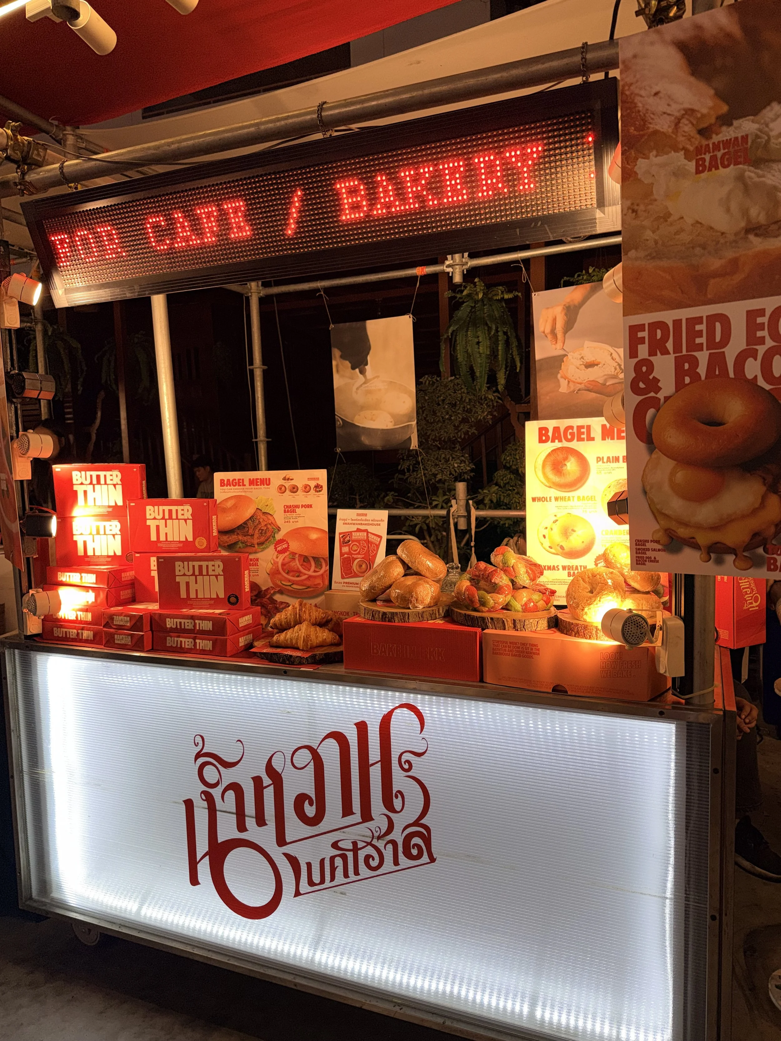 A food cart with baked goods, fried egg and bacon, and packaged butter, with menus and advertisements displayed, illuminated by warm lighting.