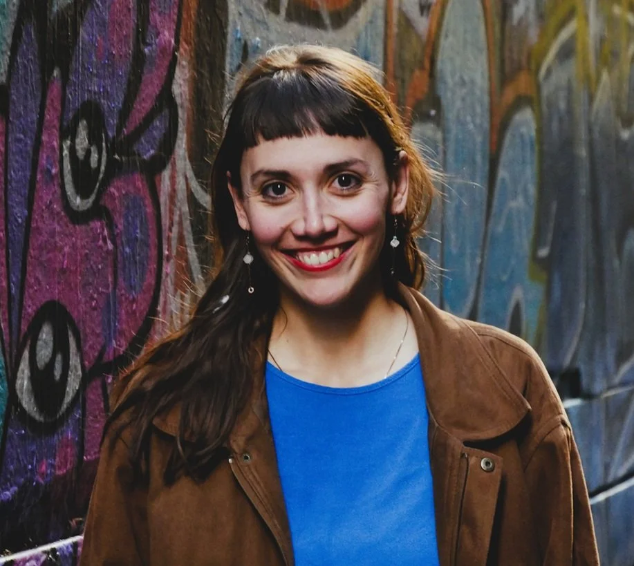 eine junge Frau mit Pony und brauner Jacke steht lächelnd vor einer Graffiti Wand