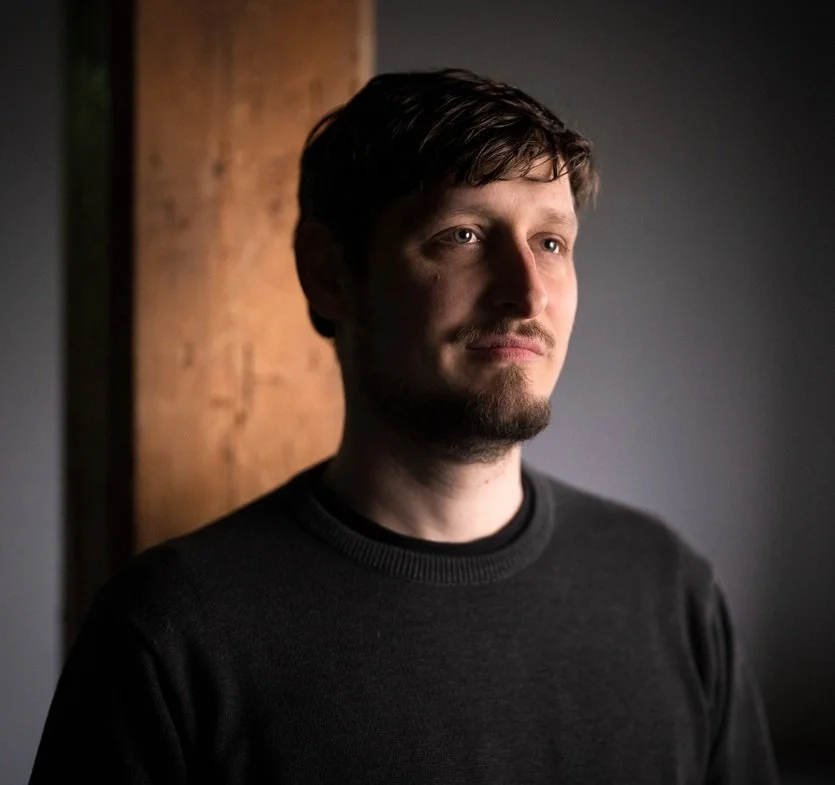ein Mann mit dunkelbraunen Haaren und Bart steht vor einem grauen Hintergrund. es ist eine Portraitaufnahme
