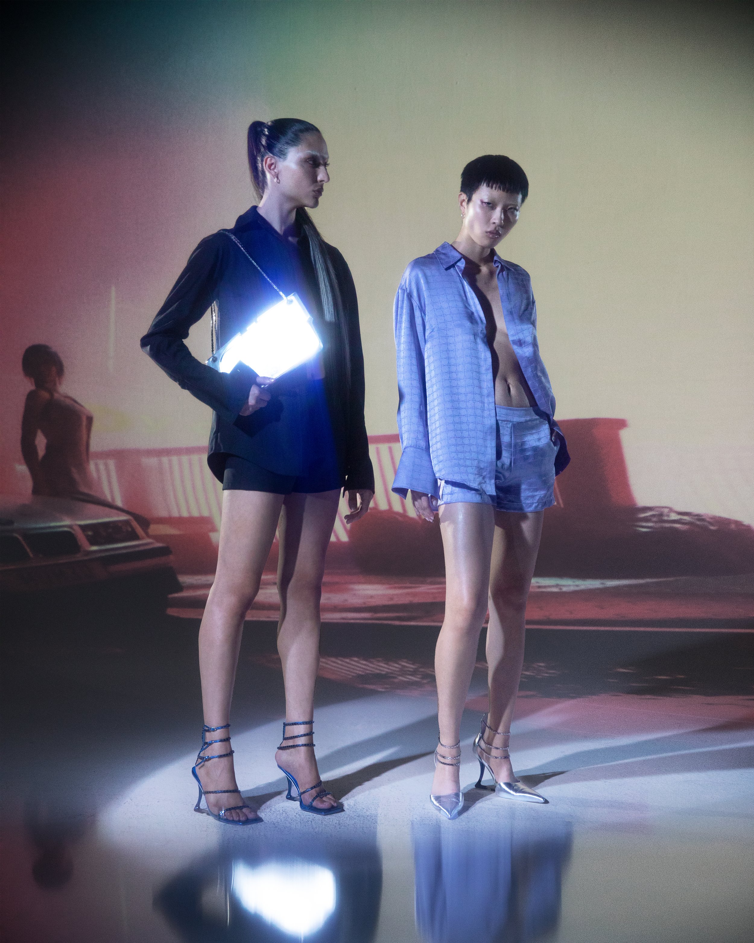 Two models standing in a fashion show wearing stylish outfits, one in a black ensemble holding a reflective bag, and the other in a blue satin set. Background features a car and neon lighting.