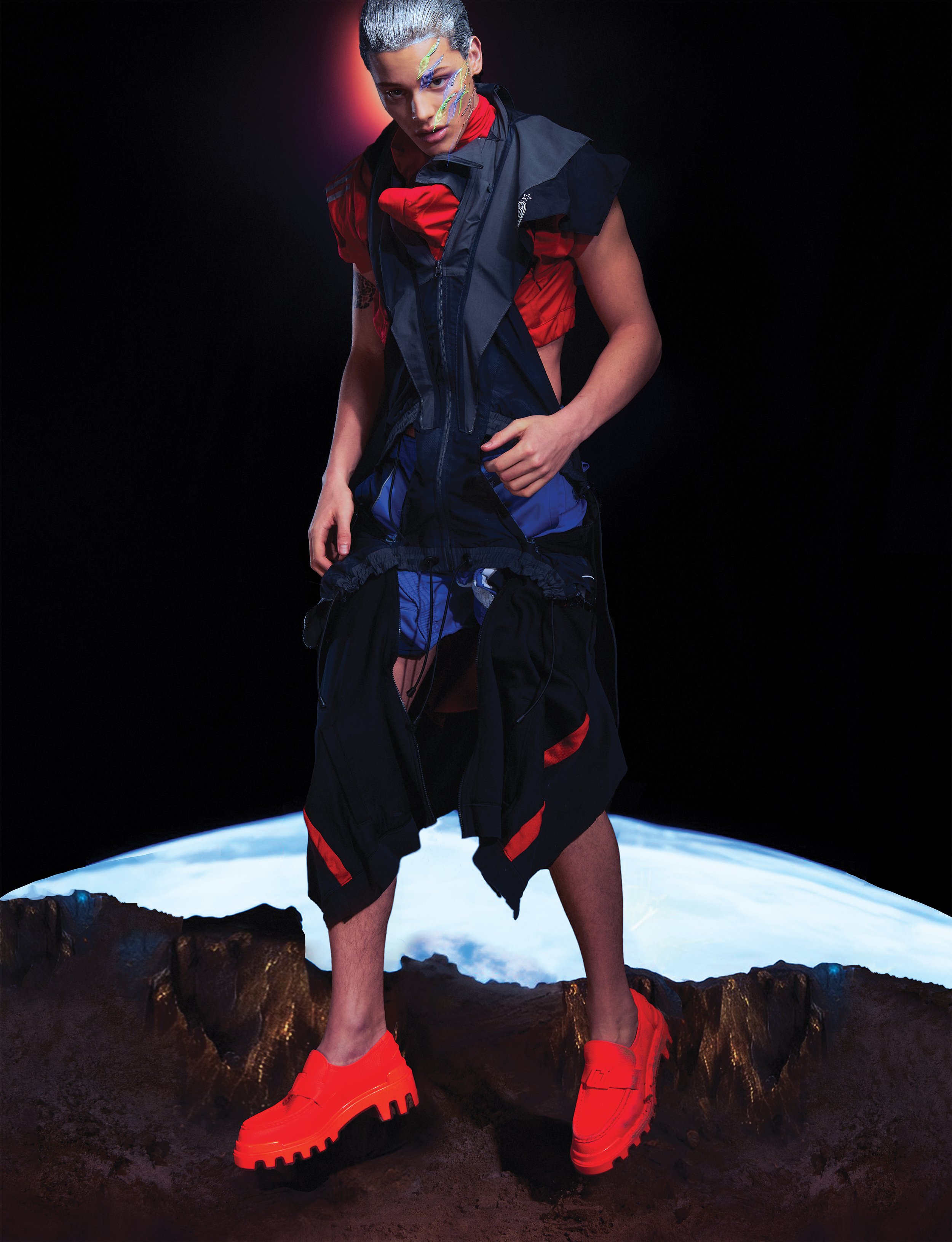 A person wearing a colorful, avant-garde outfit with a reflective vest and bright orange shoes, standing against a dark background with an illustration of Earth.