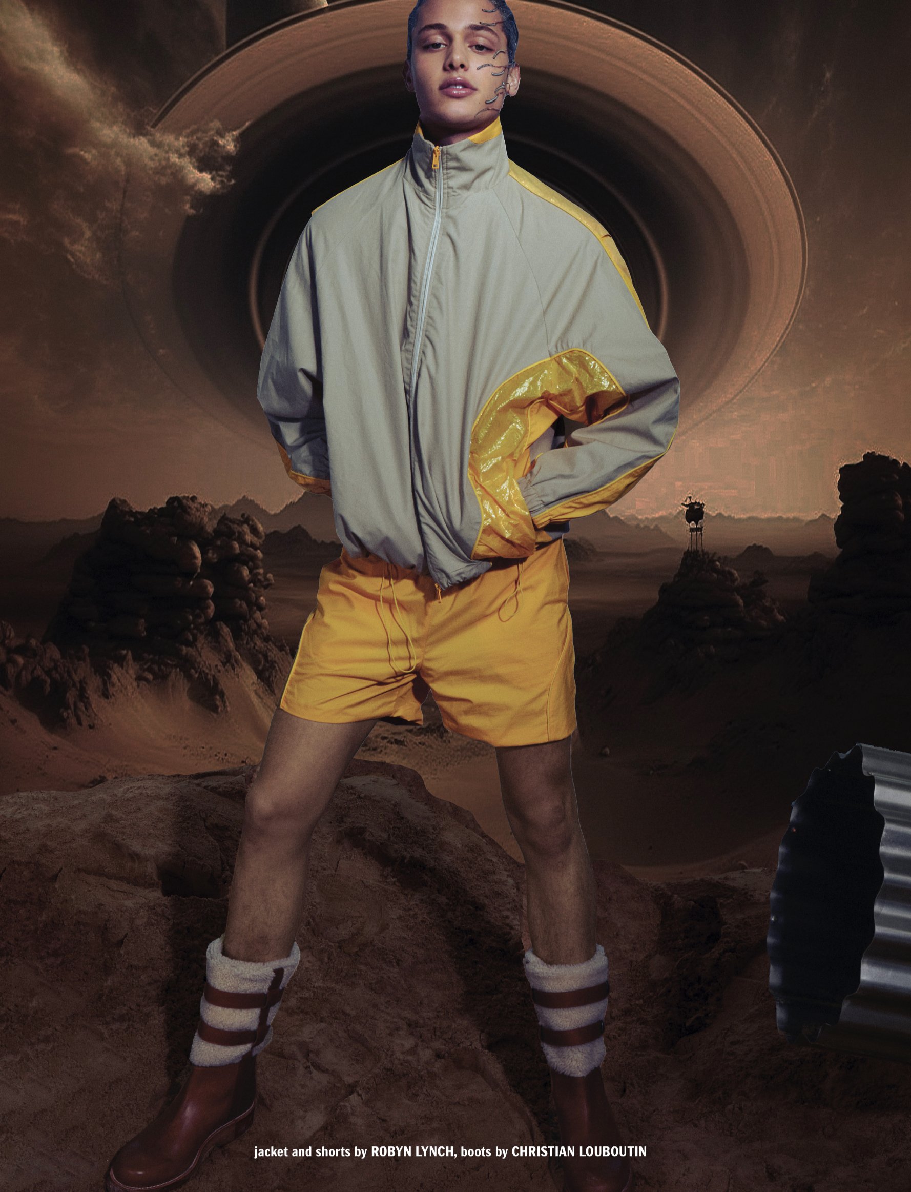Person wearing a gray and yellow jacket, yellow shorts, and brown boots with a furry trim, standing on a rocky terrain with a large ringed planet in the background. Fashion credits: Jacket and shorts by Robyn Lynch, boots by Christian Louboutin.