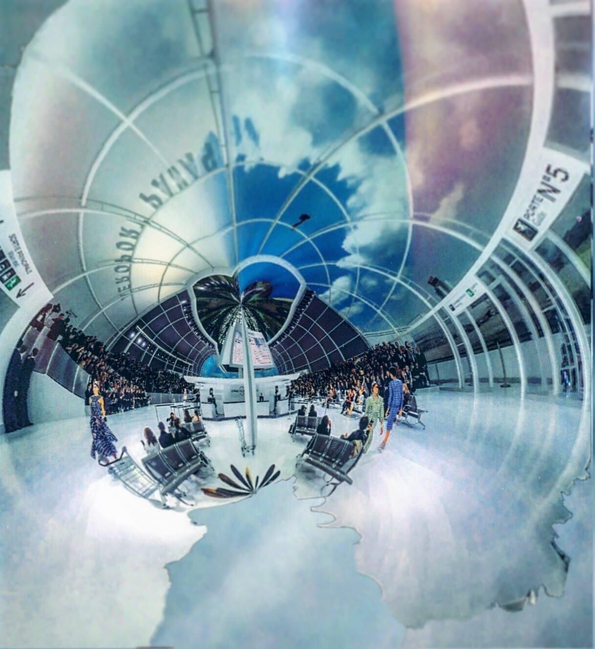 Distorted image of a large modern indoor space with a curved, glass ceiling, blue sky reflection, and people seated and standing around the edges.