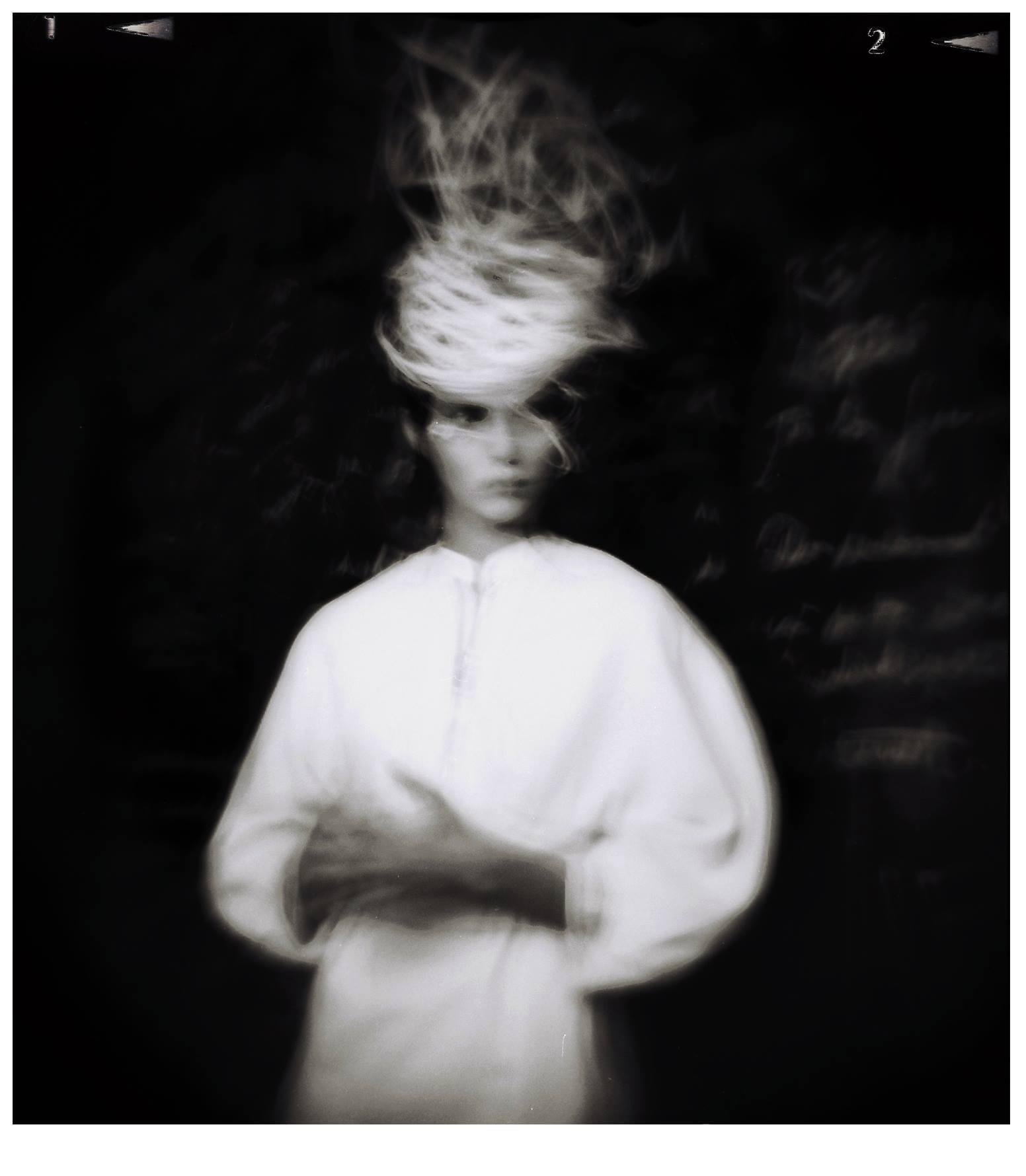 Blurry black and white image of a person with a distorted face and abstract hair swirl, wearing a white outfit against a dark background.