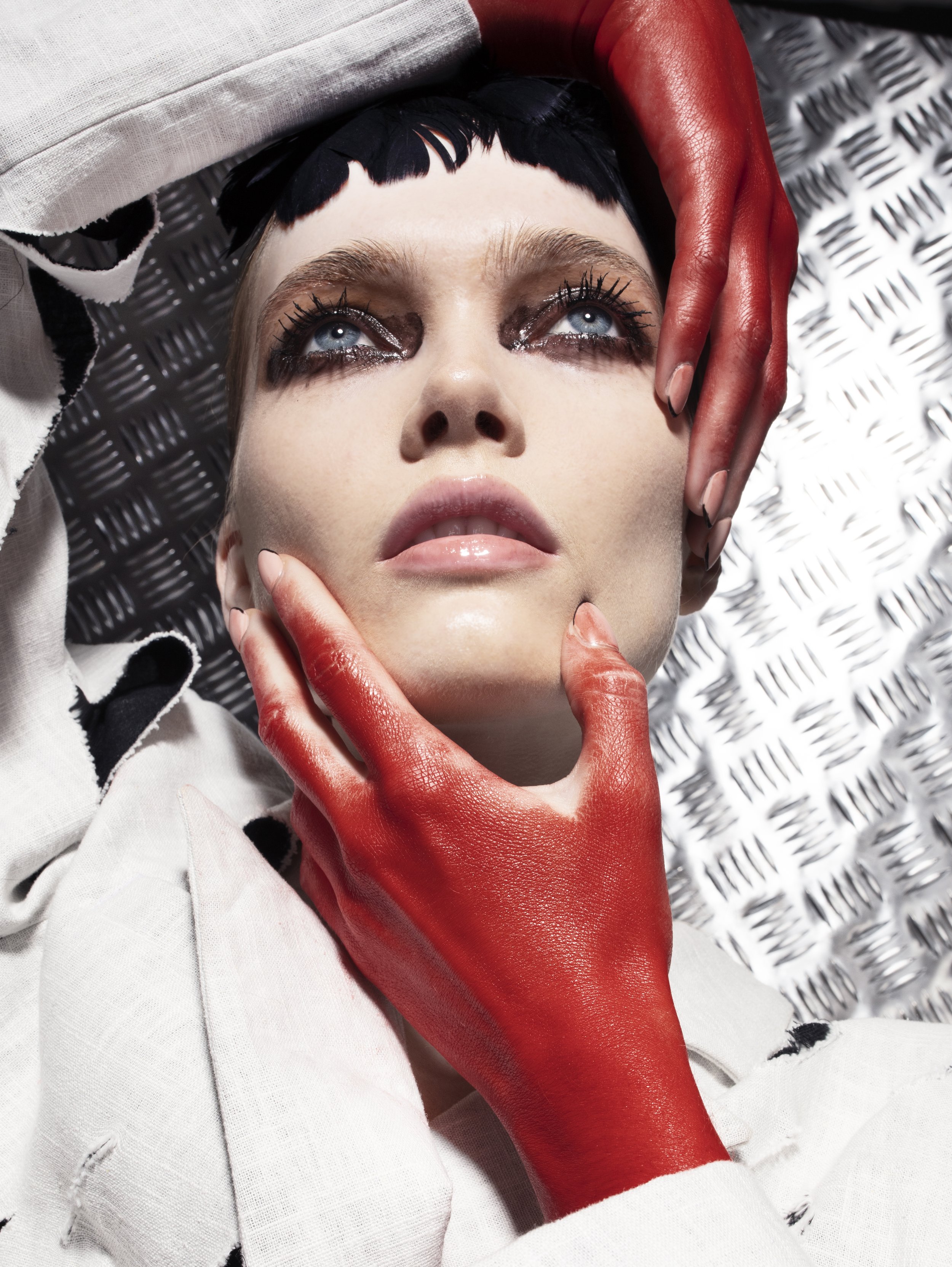 Close-up of a woman's face with dramatic eye makeup, black hair, and red-painted hands touching her face against a textured metallic background.