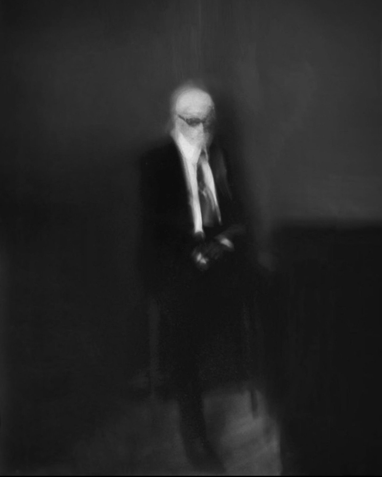 Blurred image of a person in a suit and tie, sitting on a chair against a dark background. The facial features are indistinct.
