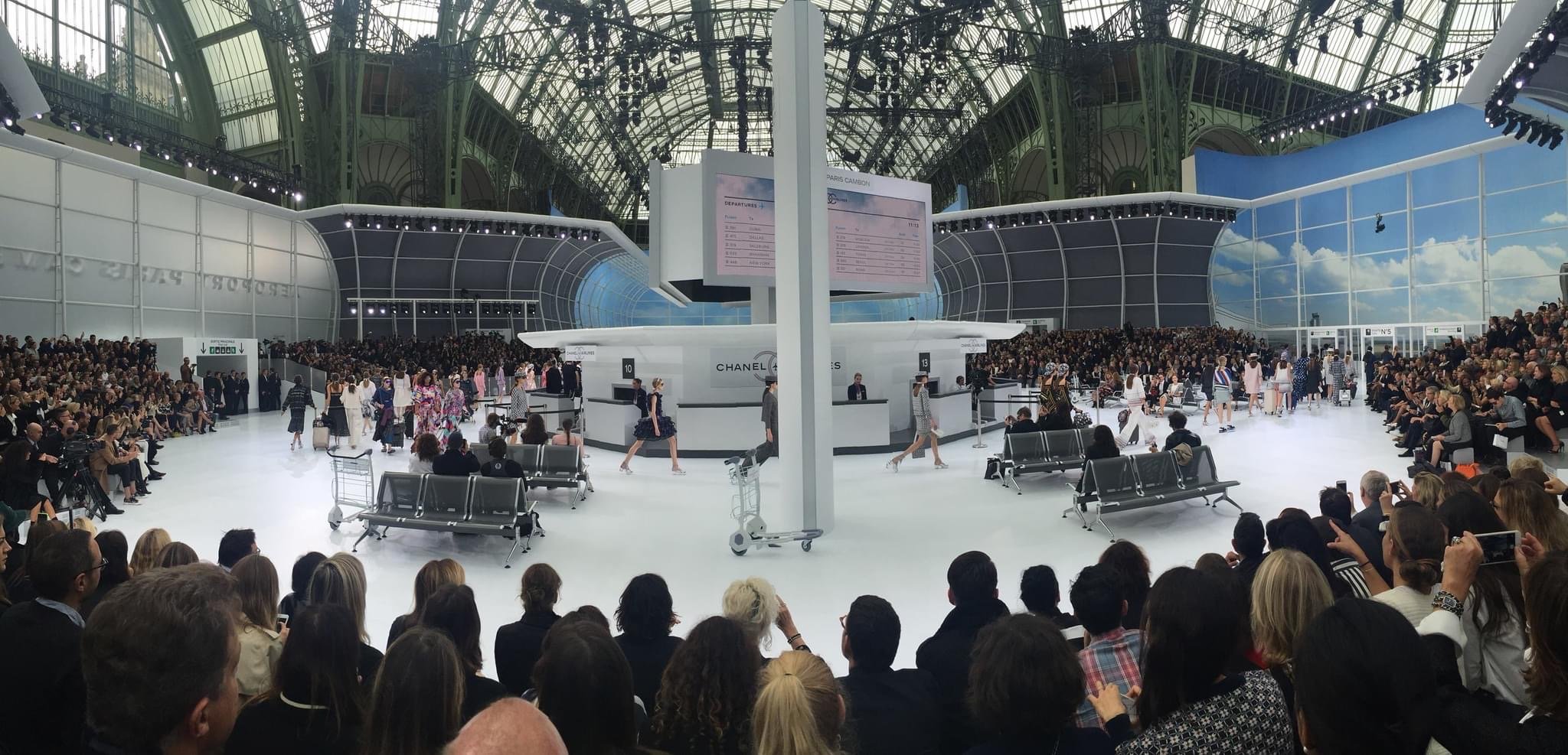 Fashion show at an indoor venue resembling an airport terminal, featuring models walking on a runway bordered by large crowds. The ceiling has a glass and metal structure.