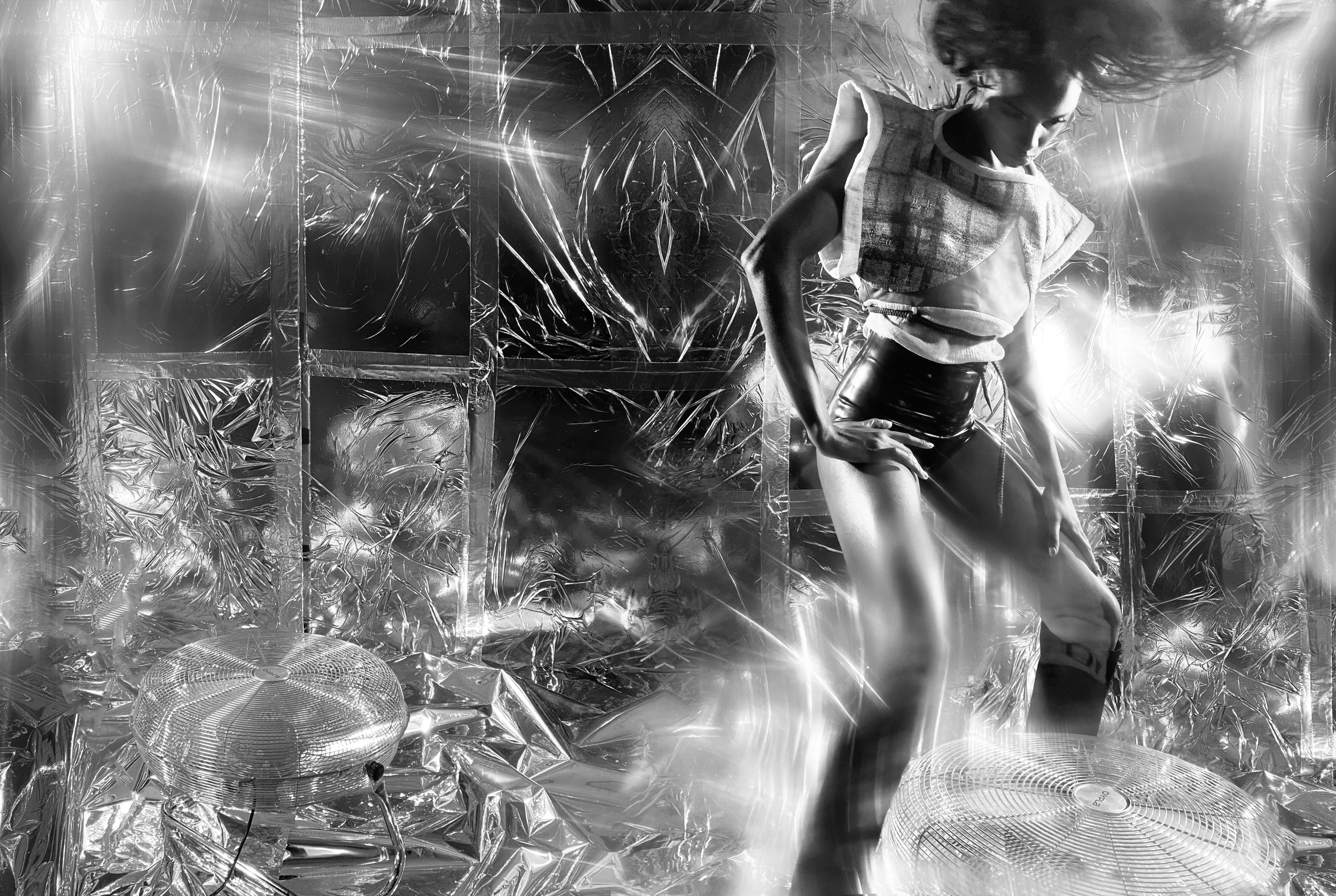 Black and white image of a person dancing energetically in a room covered with reflective foil material. There are two electric fans on the floor, creating movement in the scene.