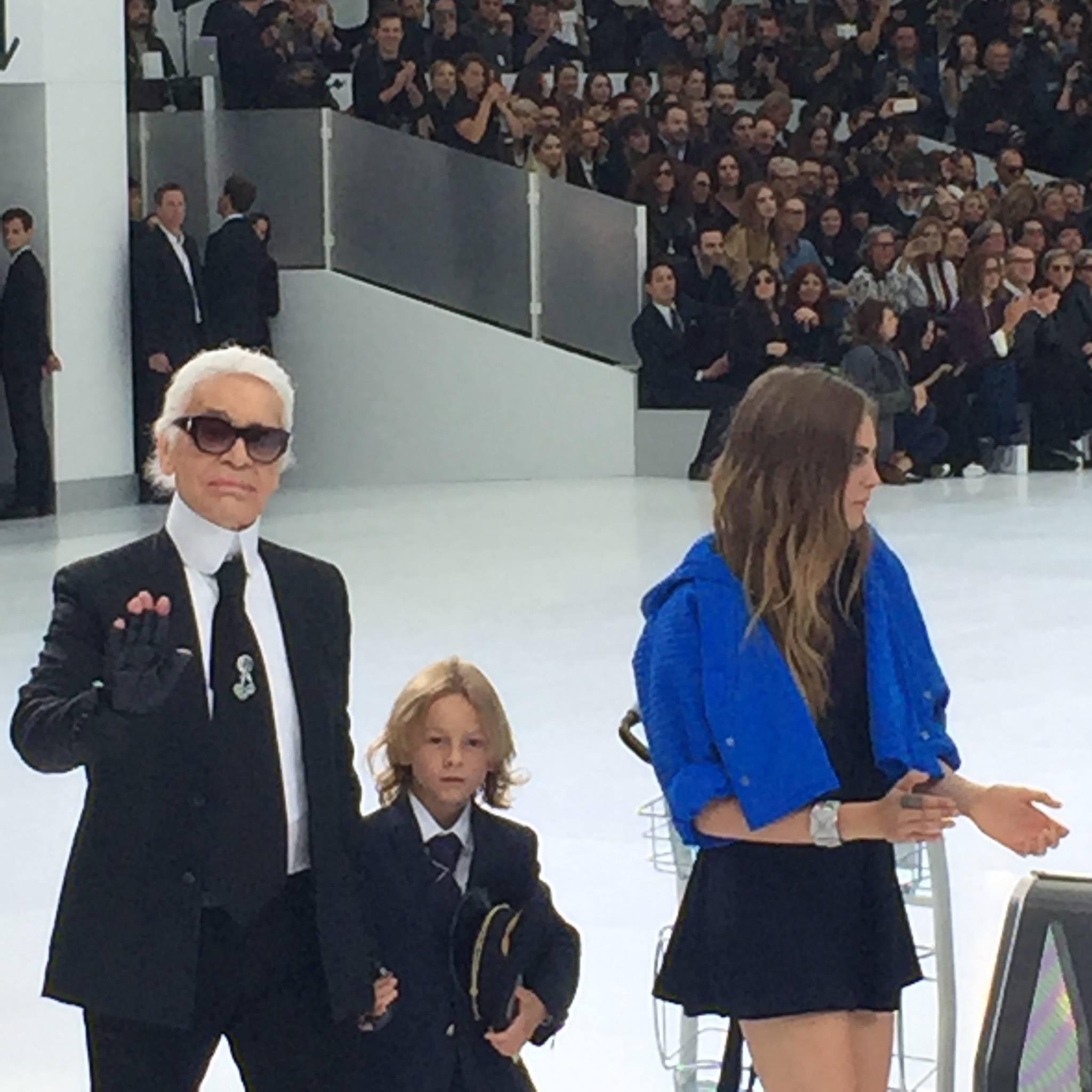 Fashion show with a man in sunglasses and suit, a child in a suit, and a woman in a blue jacket and black dress on the runway, with an audience watching.