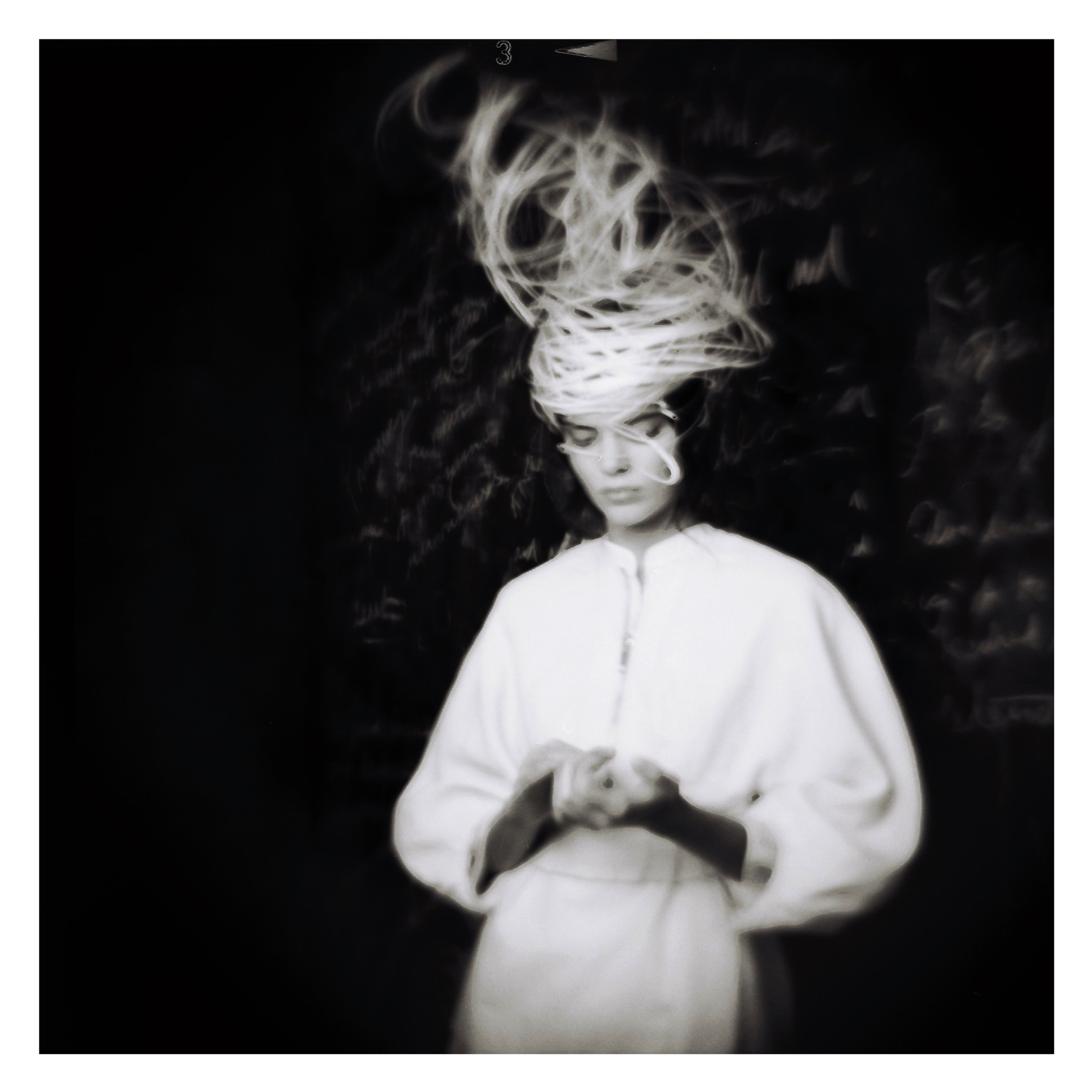 Black and white photo of a person in a white outfit, with intricate, artistic headpiece. The background is dark with blurred writing.