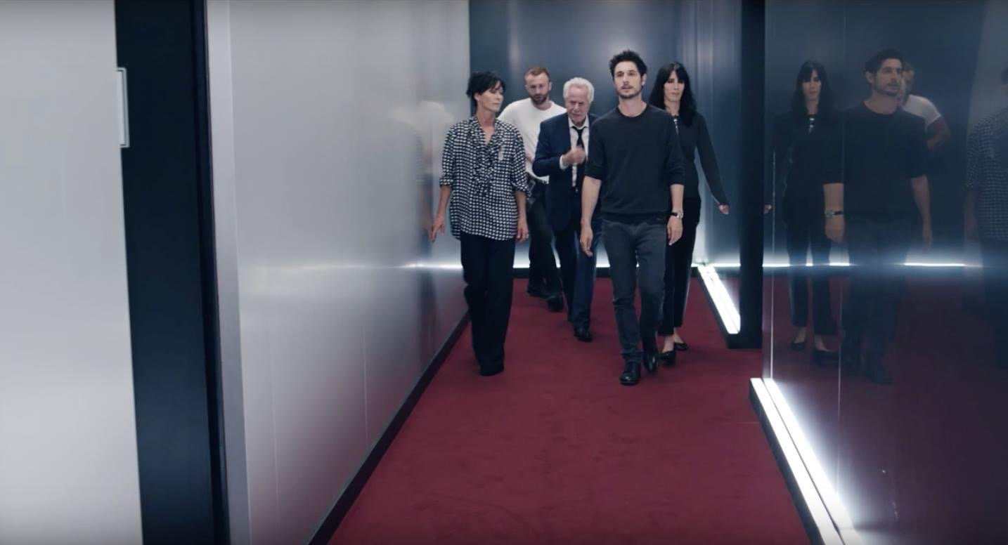 A group of people walking down a narrow hallway with red carpet and mirrored walls, giving a modern and sleek appearance.