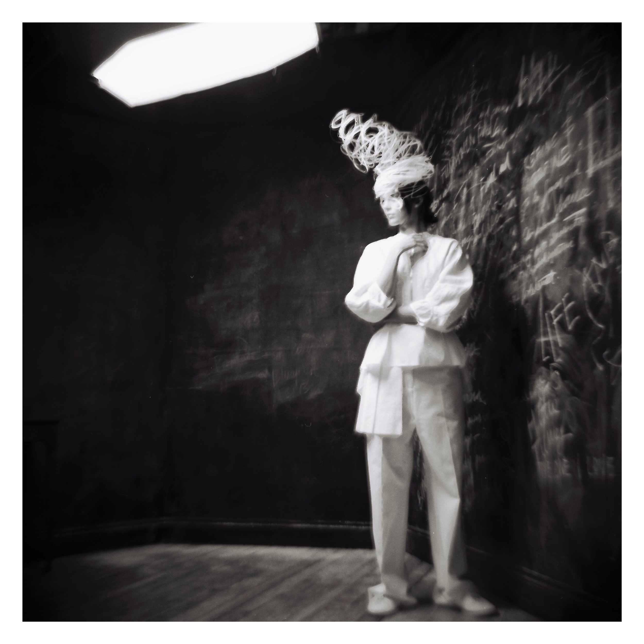 A person dressed in a white outfit and intricate headpiece stands in a dimly lit room with chalkboard walls covered in writing. The image has a blurred, artistic effect.