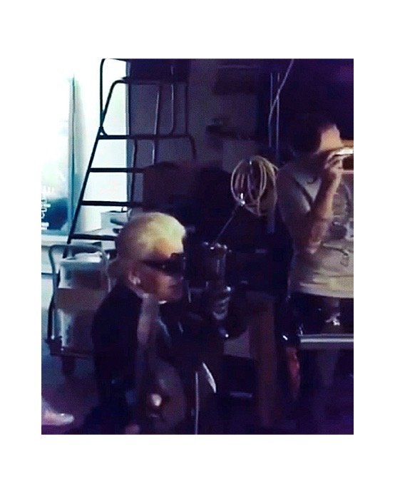 Person with short blonde hair and sunglasses holding a device, standing in a cluttered room with another person eating.