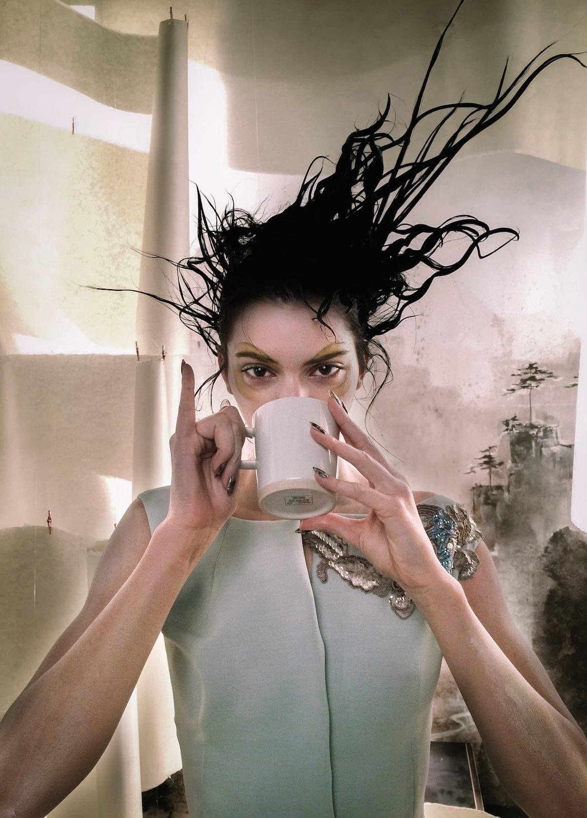 A person with stylized hair holds a white mug at their mouth, wearing a light-colored outfit against an abstract background.