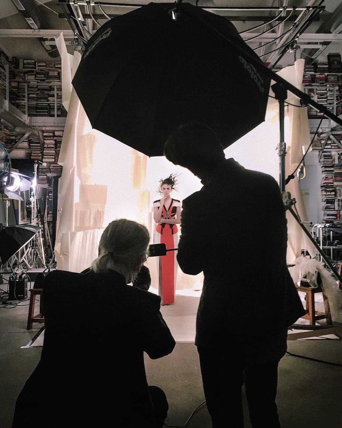 Photo shoot in studio with model, photographer, and assistant under large black umbrella light.
