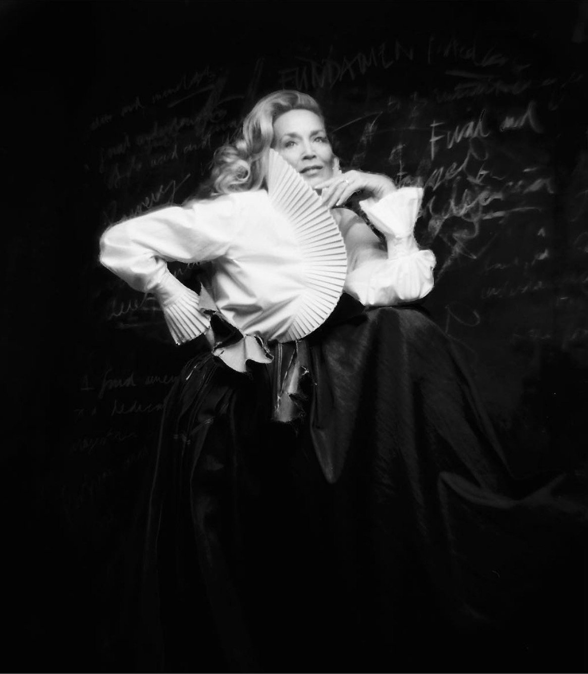 Black and white portrait of a person wearing a dramatic outfit with a high collar and pleated design against a textured backdrop.