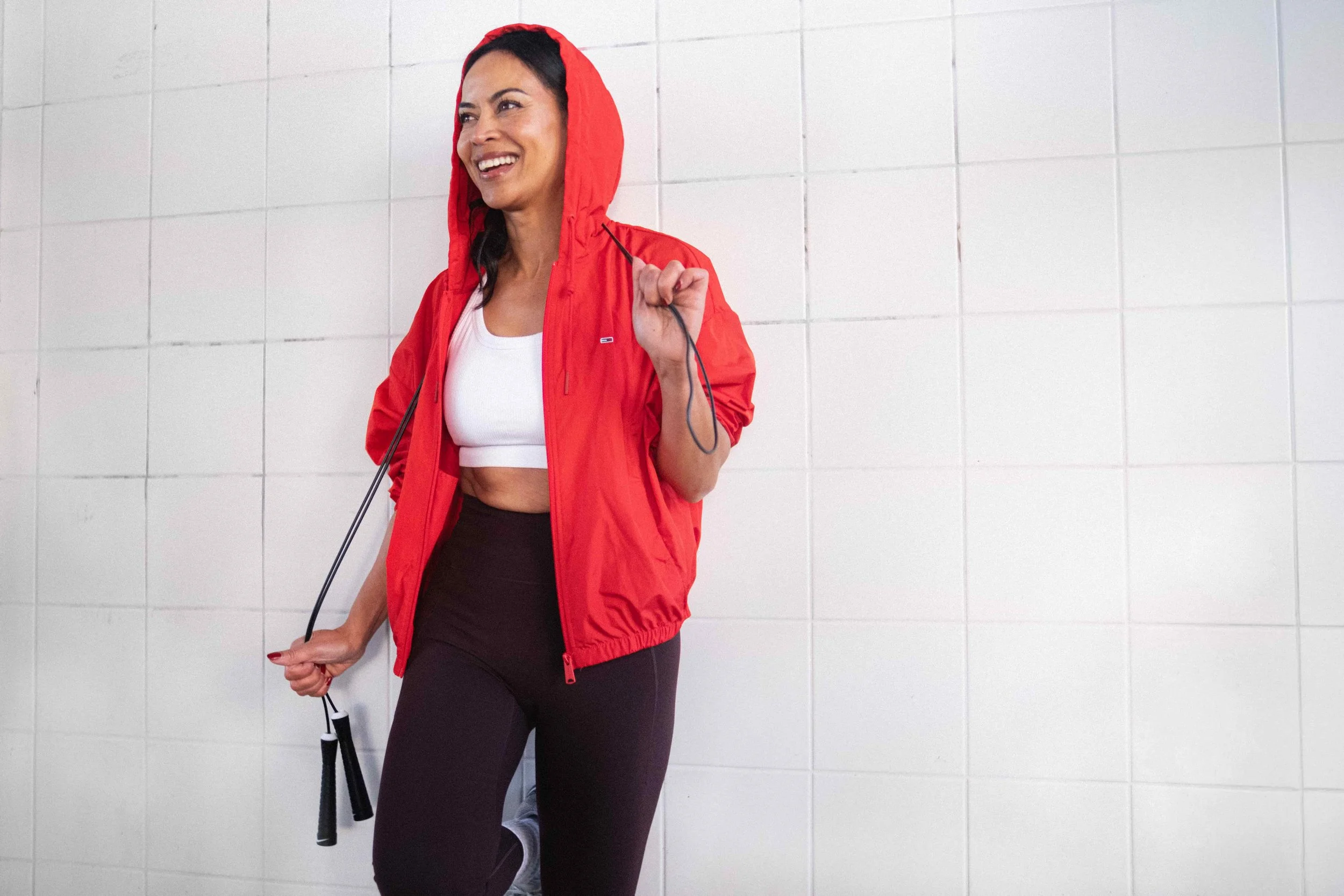 Vrouw in sportkleding met rode jas en springtouw, lachend, staand tegen een witte tegelmuur.