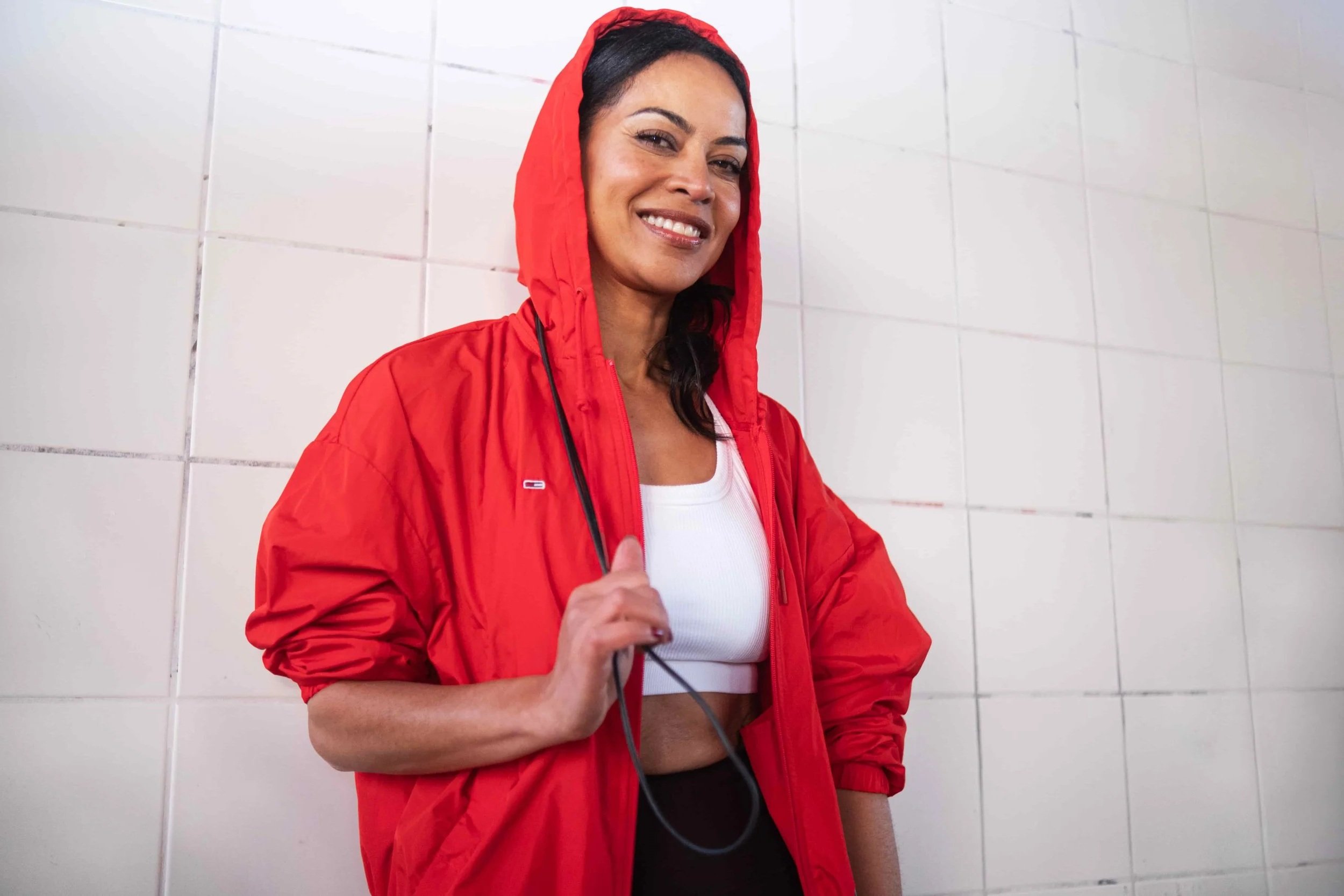 Vrouw met een rode jas en hoodie, glimlachend naast een witte tegelmuur.