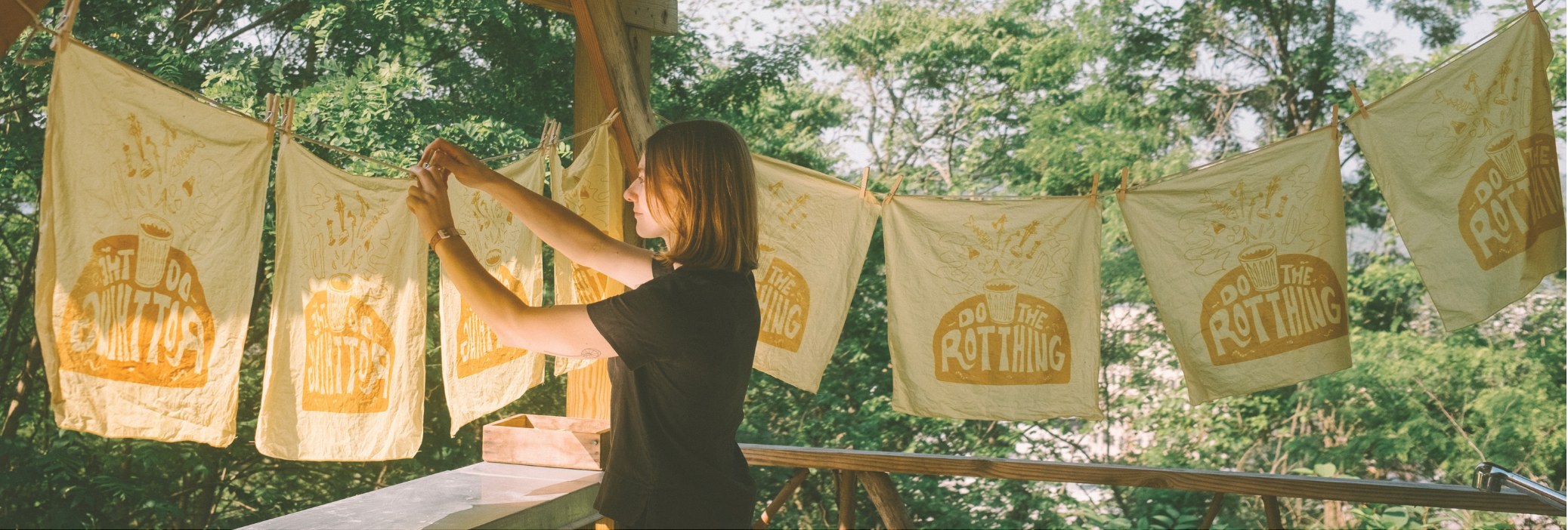 Hannah hangs bandanas printed with marigold dye on a clothesline outdoors with a green forested background
