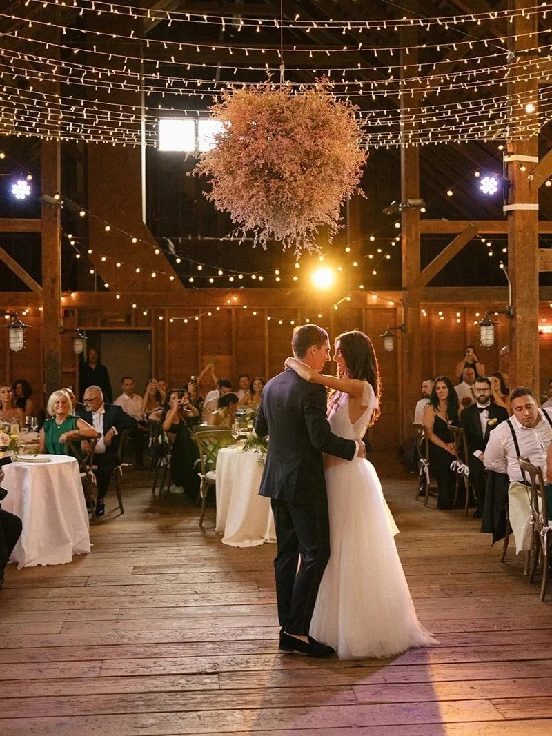 A couple is dancing in the center of a warmly lit rustic wedding reception with string lights and hanging decorations, while seated guests watch and take photos.
