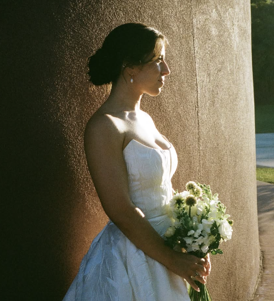 A woman in a white wedding dress holding a bouquet of white flowers, standing against a textured brown wall in sunlight.