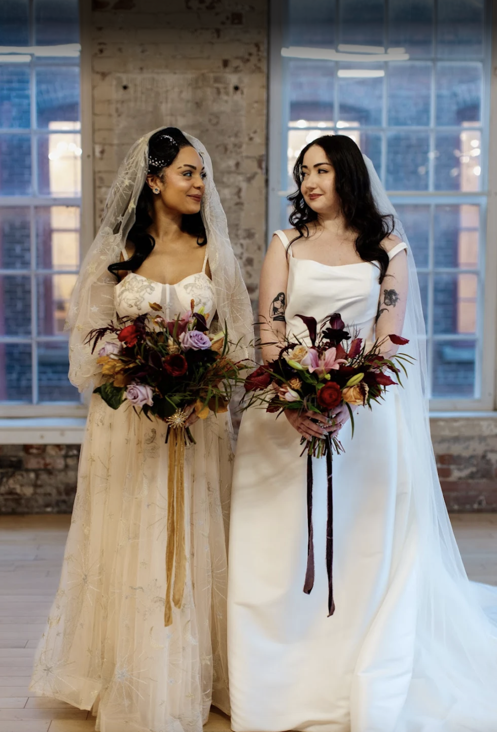 Two women in wedding dresses holding bouquets, standing in a bright room with brick and large windows, looking at each other.