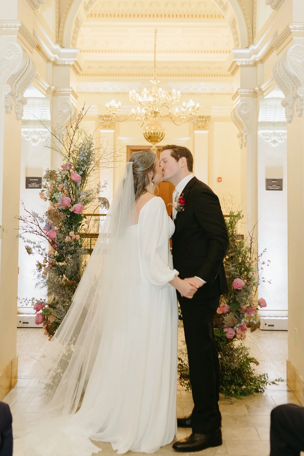 Bride and groom kissing at their wedding ceremony, surrounded by floral arrangements in a decorated indoor venue with chandeliers and ornate architectural details.