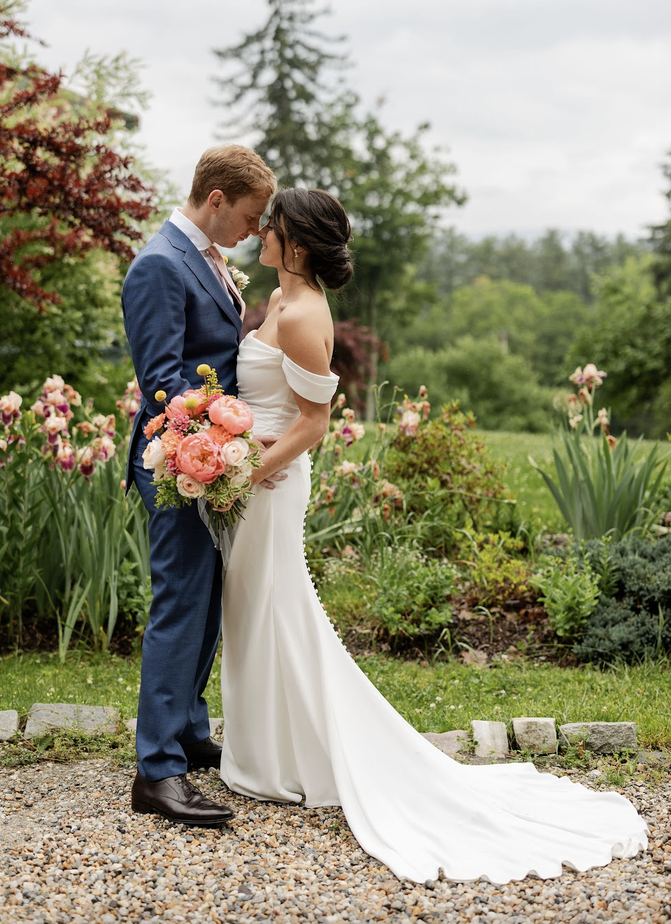 A bride and groom standing close with foreheads touching, outdoors in a garden, with the bride holding a colorful bouquet of flowers.