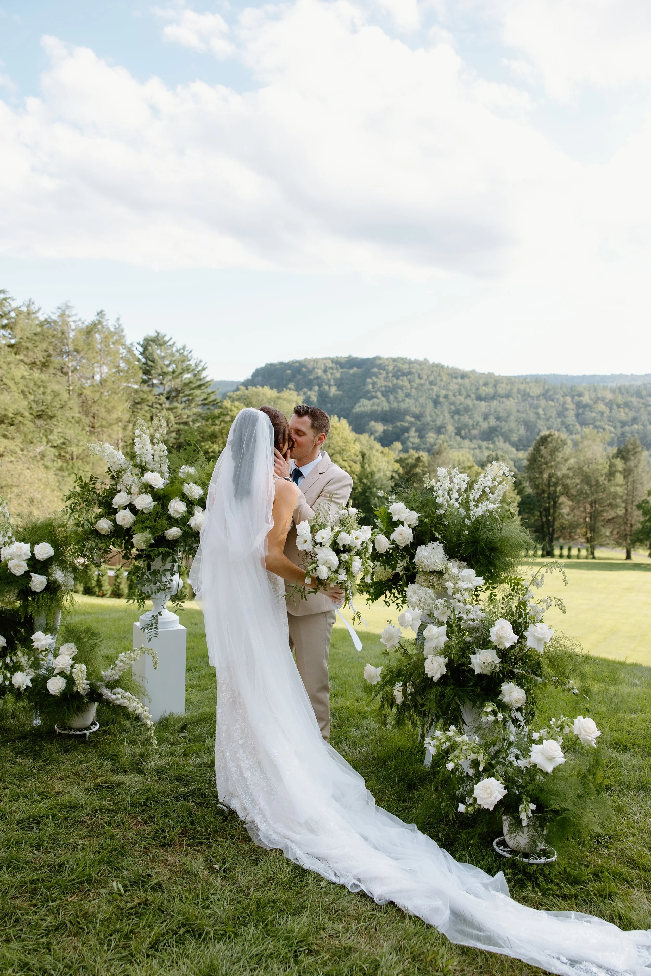 A bride and groom kiss at an outdoor wedding ceremony decorated with white flowers and greenery, set against a scenic landscape of green hills and trees under a partly cloudy sky.