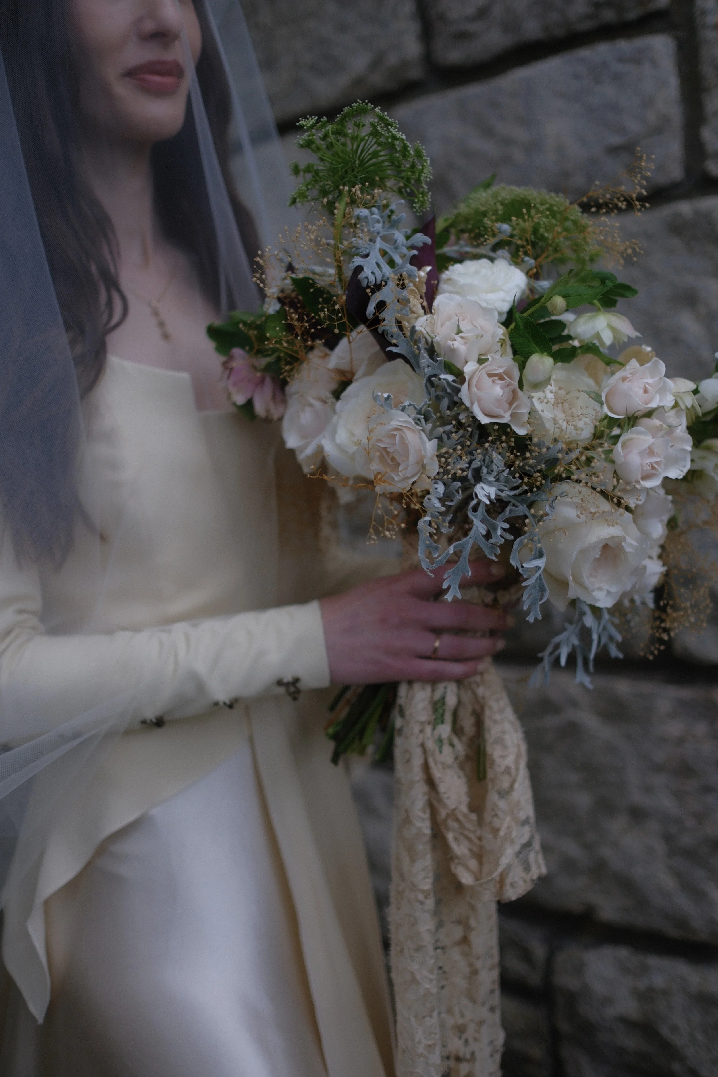 A bride holding a bouquet of white and pink flowers with greenery, velvet, and lace ribbons, standing against a stone wall.