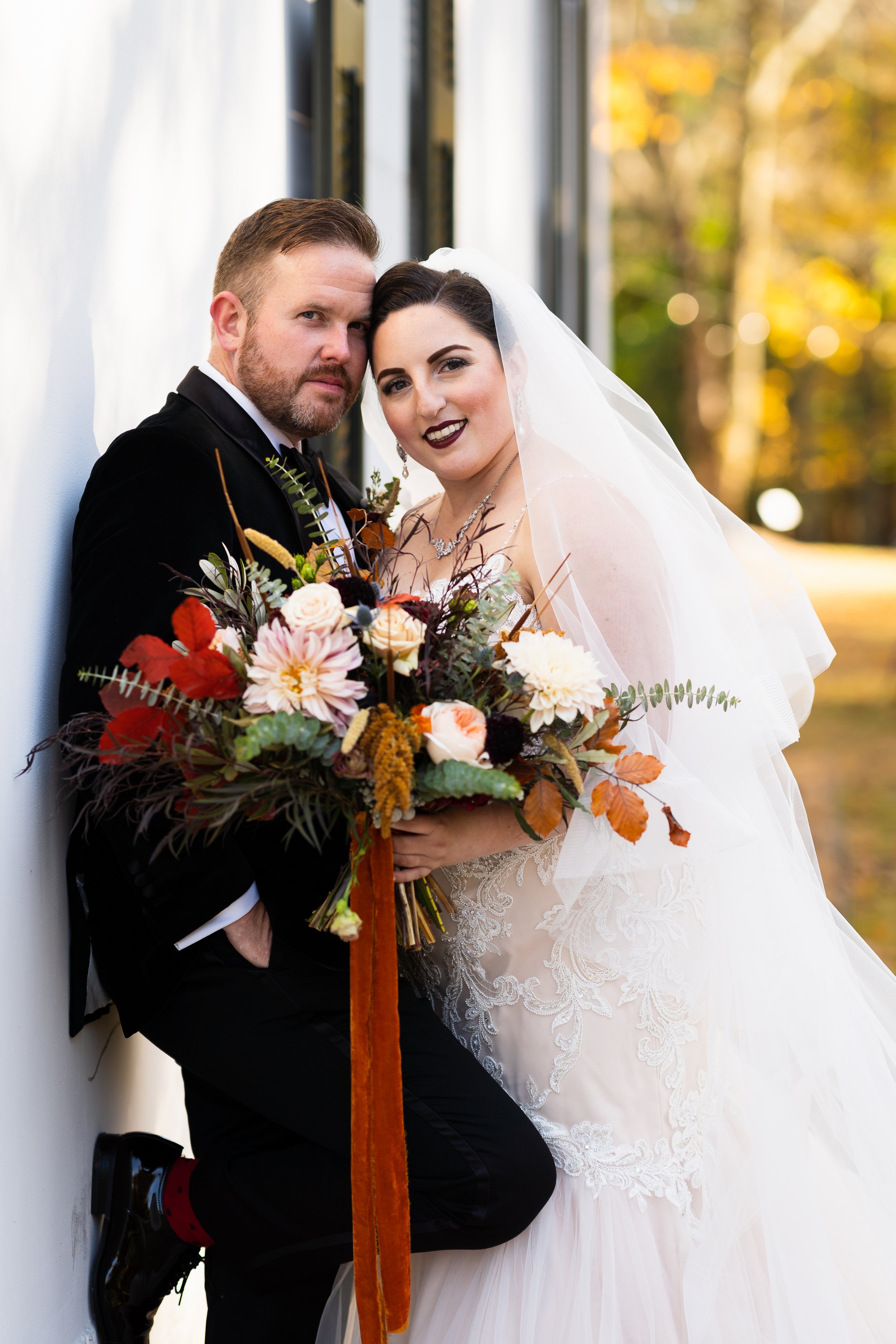 A bride and groom in wedding attire standing close together outdoors, holding a bouquet of flowers, with trees and autumn leaves in the background.