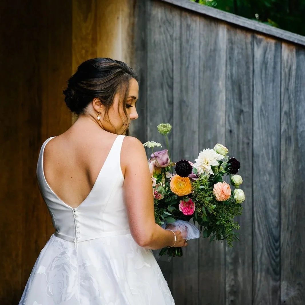 Bride with brown hair in a white wedding dress holding a colorful bouquet, standing next to a wooden fence.