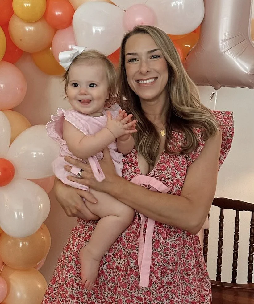 A woman holding a smiling young girl at a celebration with pink, peach, and orange balloons in the background.