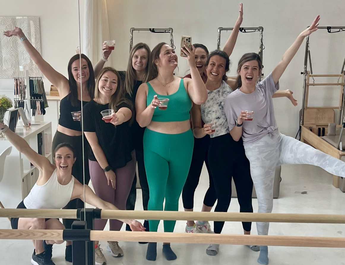 Group of eight women in workout attire celebrating with drinks and taking a selfie in a fitness studio.